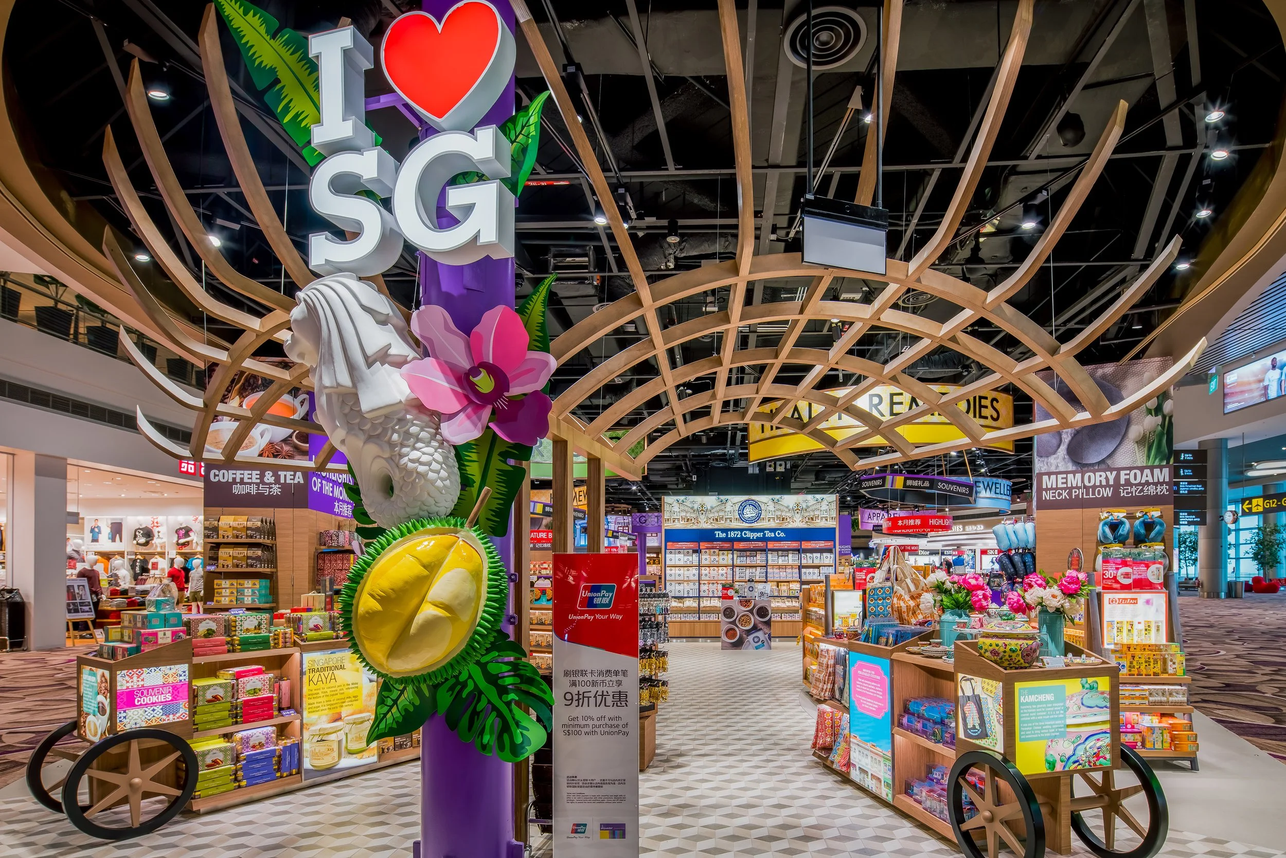 Shopping area in Singapore Changi Airport with colorful sign that reads "I ❤️ SG" decorated with flowers and leaves, surrounded by souvenir stalls and signs for various stores and services.