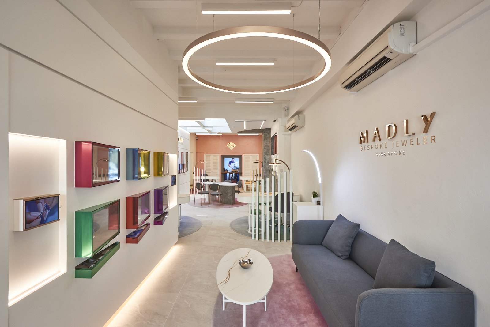 Interior view of Madly Bespoke Jeweler in Singapore, featuring a modern, minimalist design with a gray sofa, colorful display frames on the wall, a white round coffee table, and a circular ceiling light fixture.