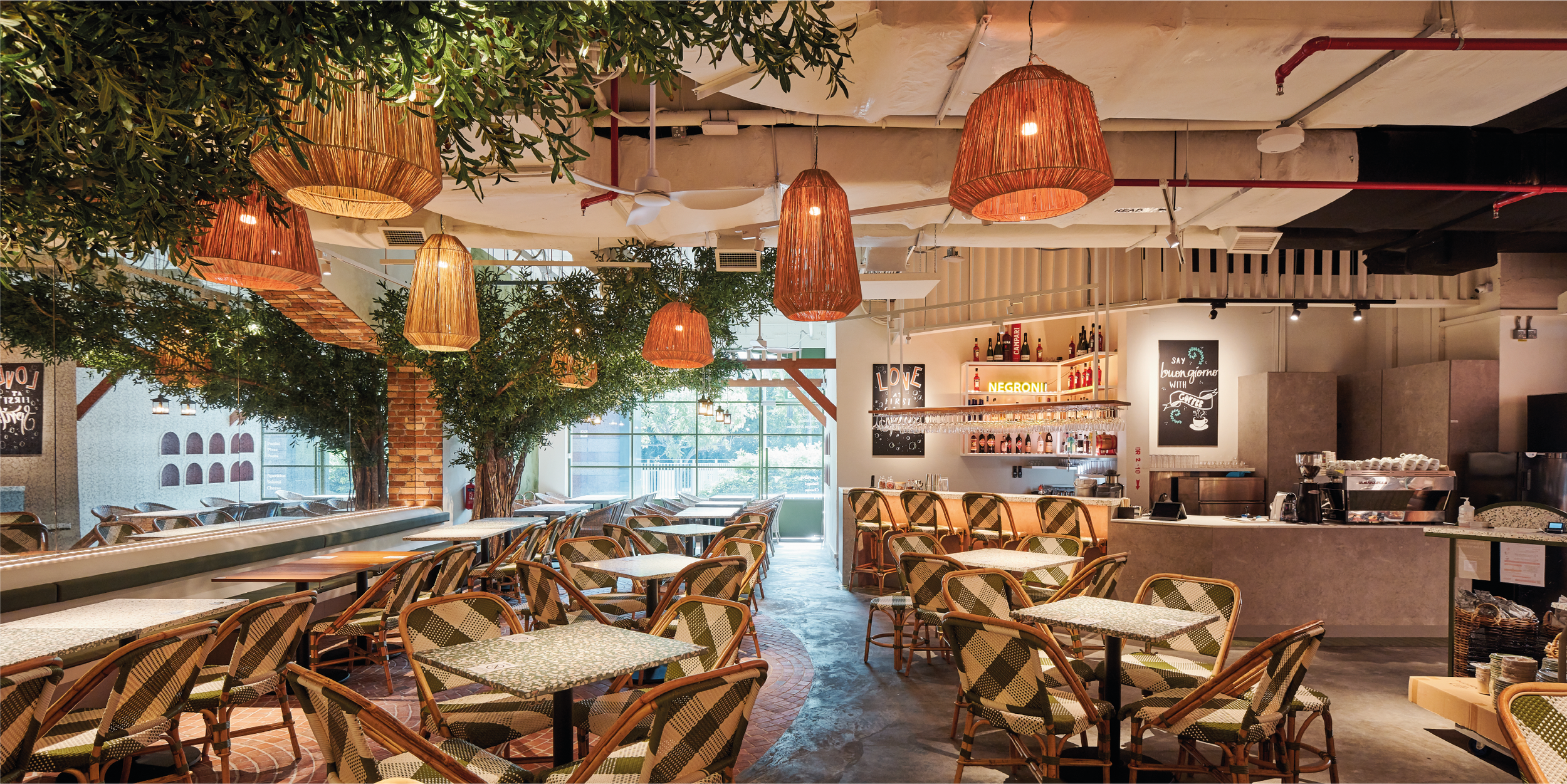 Empty restaurant with green trees and hanging woven lamps, wooden chairs with patterned cushions, a bar with bottles, and large windows letting in natural light.