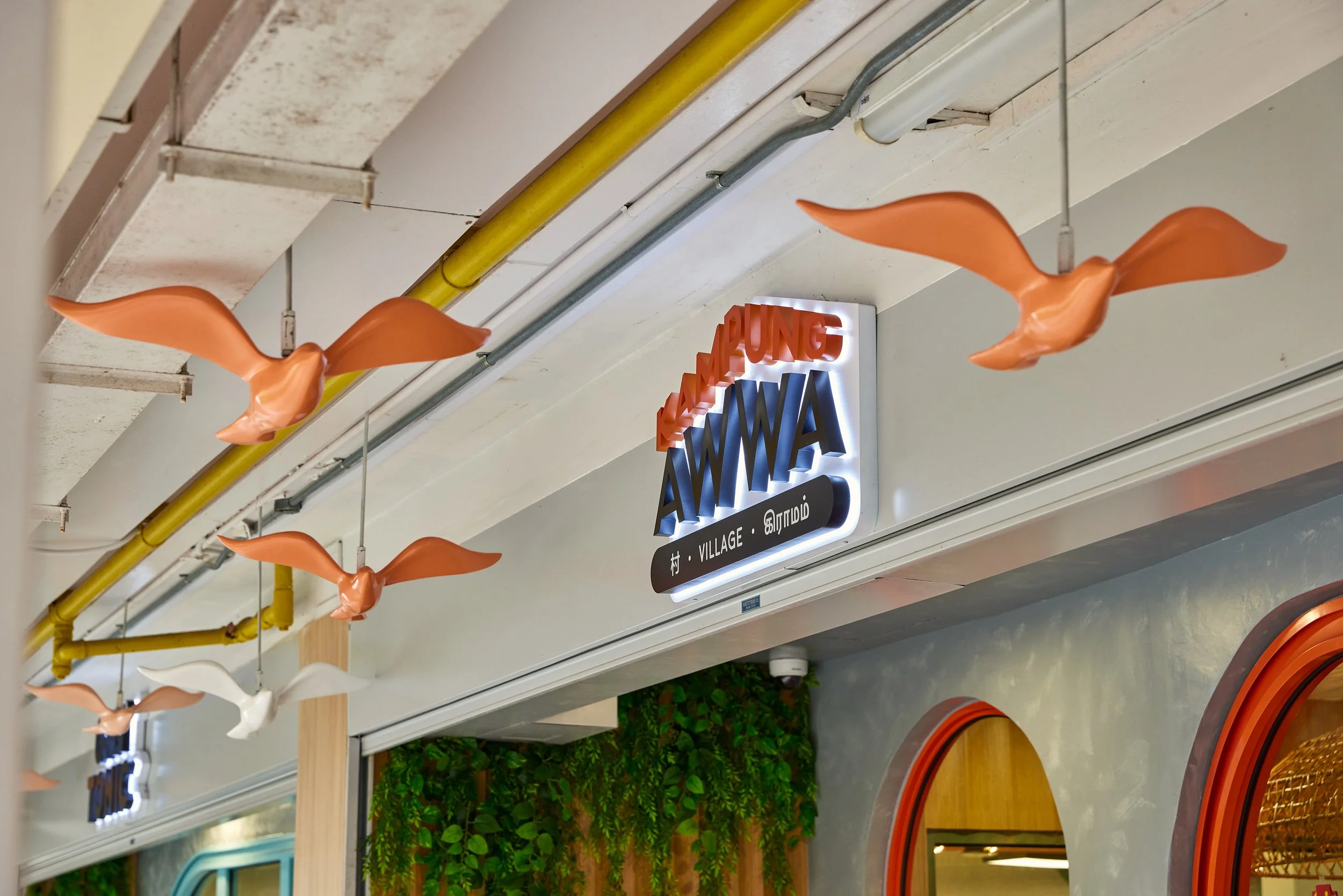 Decorative ceiling with orange bird and white bird hanging, illuminated sign reading 'KAM YUNG AWMA' and smaller sign with Asian characters, part of a storefront inside a shopping mall.