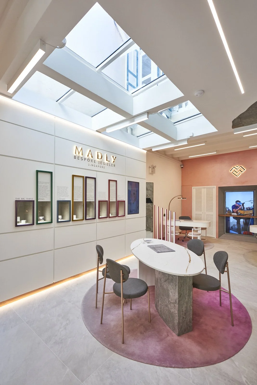 Interior of a jewelry store named MADLY, with display cases, a white marble table, four chairs, and a window showing a musician playing drums.