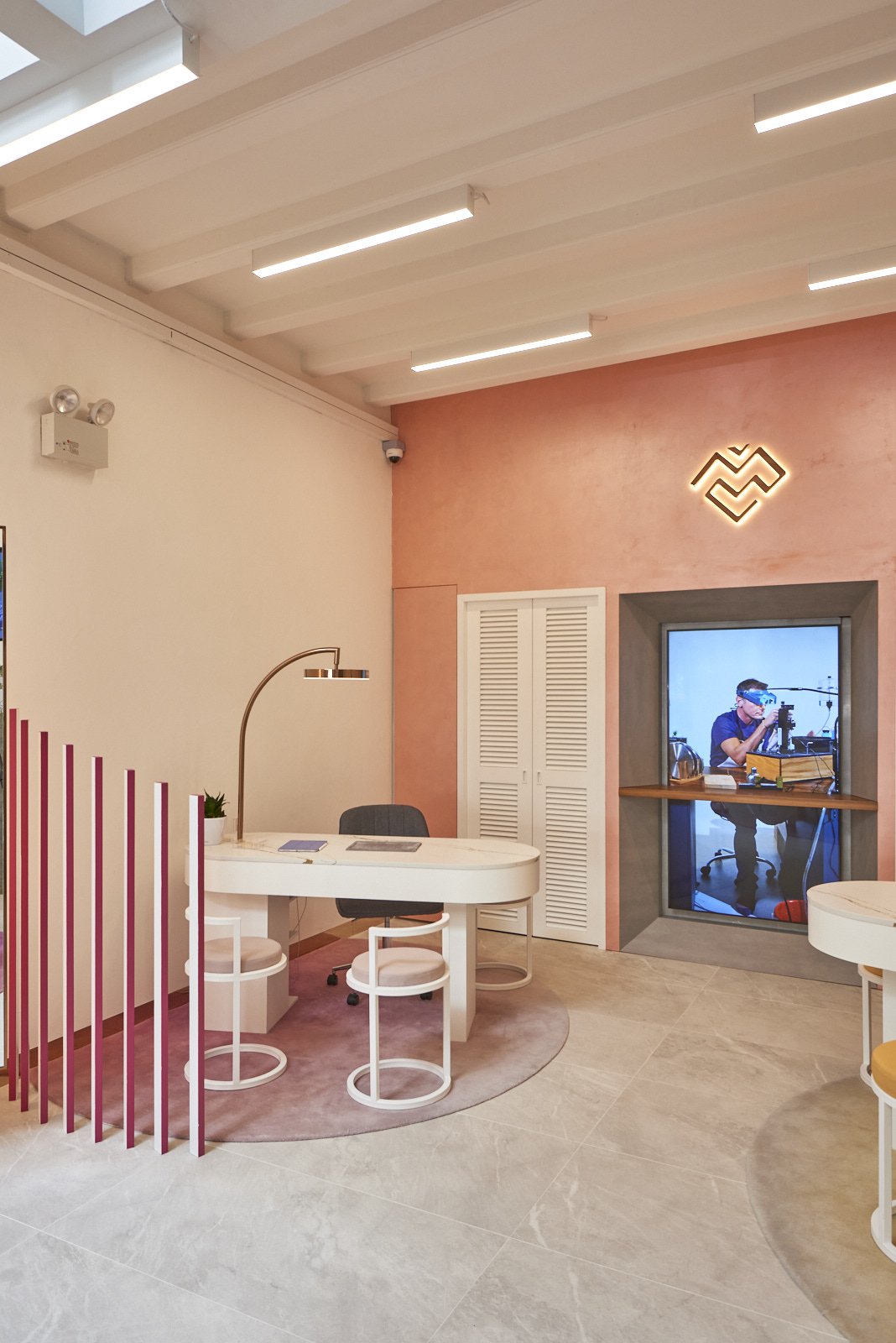 Modern office space with a white curved desk, black office chair, pink accent wall, decorative lighting, and a windowed recording booth with a person operating camera equipment.