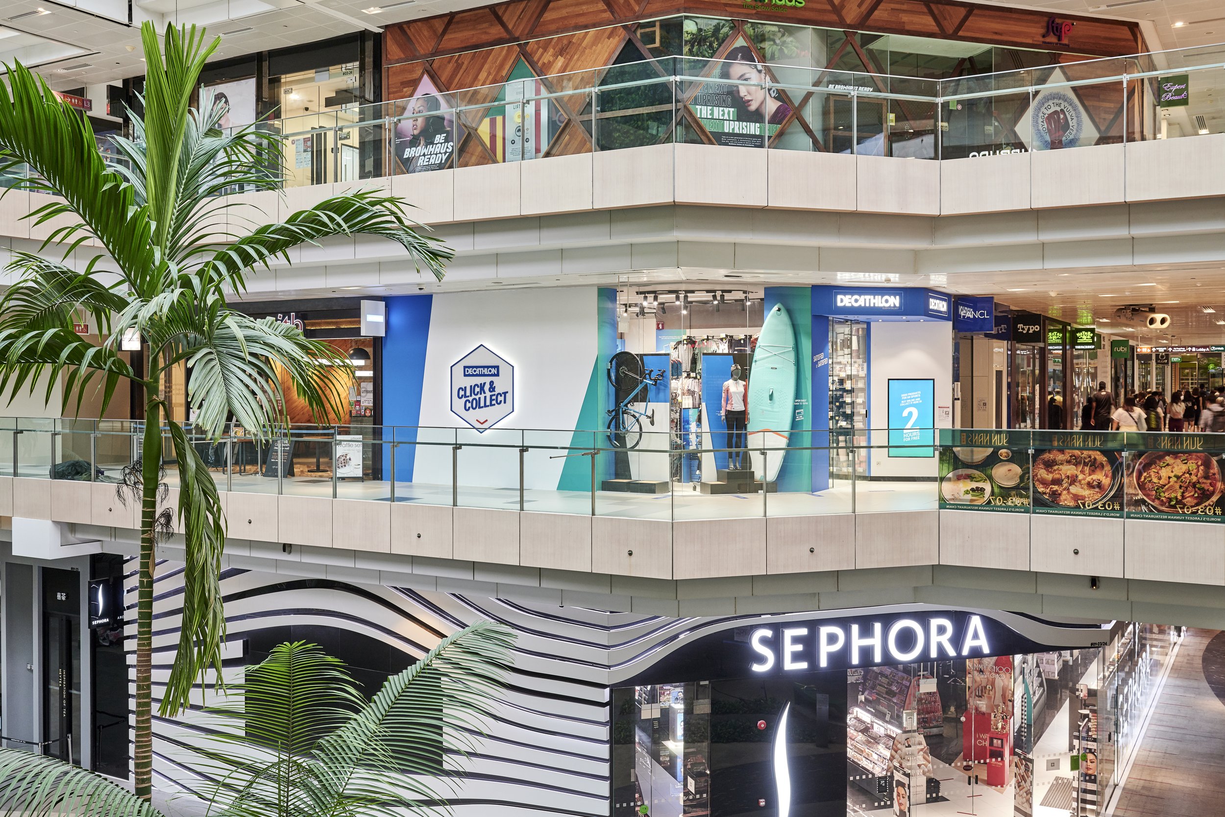 Interior view of a multi-level shopping mall featuring a Sephora store on the lower level and a Decathlon store on the upper level, with a large indoor green plant in the foreground.