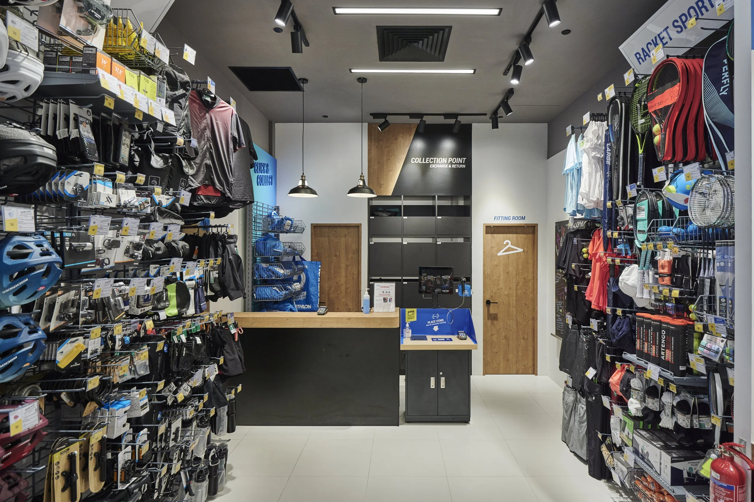 Indoor sports store checkout area with shelves of sports gear, helmets, tennis rackets, and clothing, featuring a black counter and signage for collection point, fitting room, and sports shop.