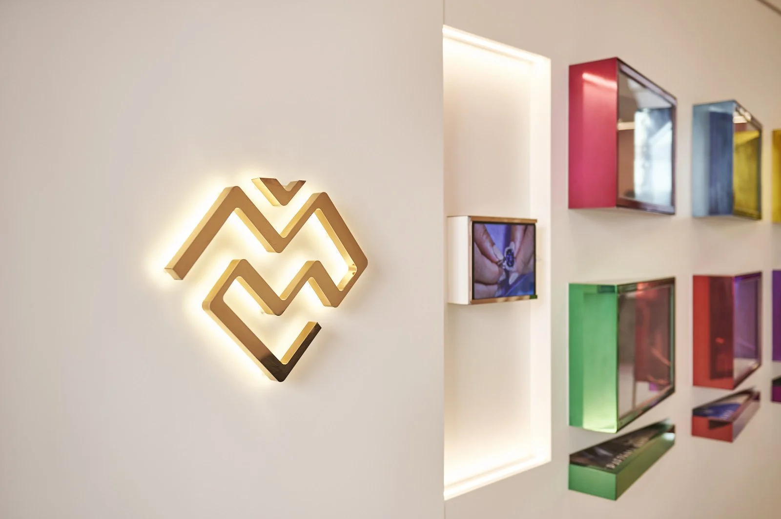 Interior of a modern building featuring a white wall with a backlit geometric logo and colorful wall-mounted display boxes.