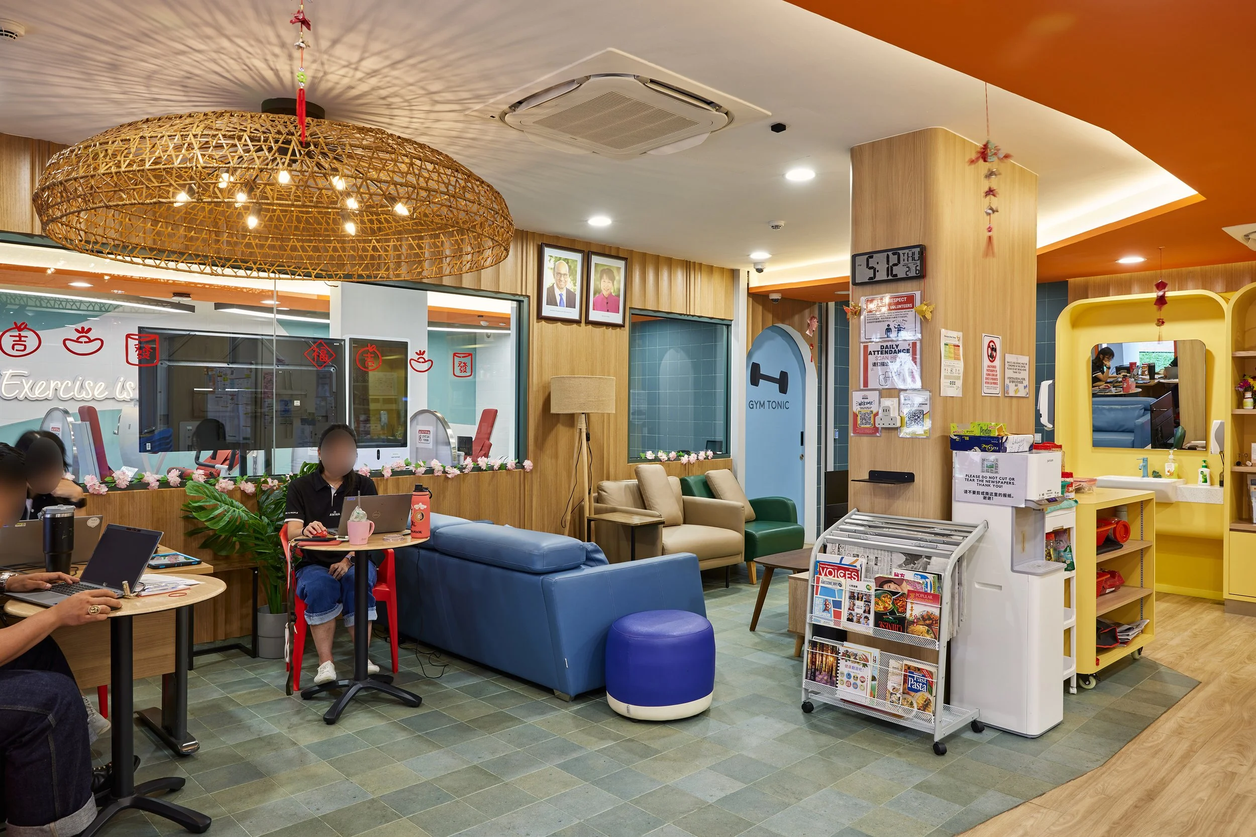 Interior of a cozy cafe or lounge area with seating, plants, and a reception desk. People are working on laptops. Decor includes framed photos, hanging ornaments, and a large woven light fixture.