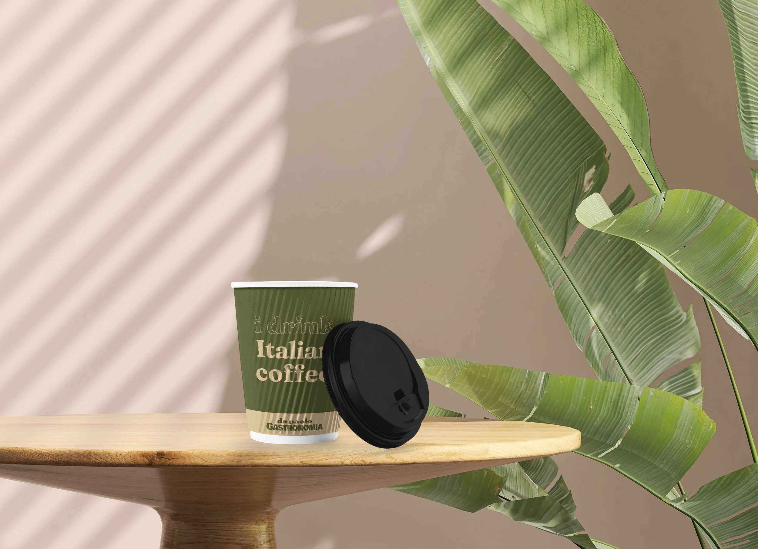 A green coffee cup with text that says 'i drink Italian coffee', a black coffee lid, on a wooden table, with a beige background and large green tropical leaves casting shadows.