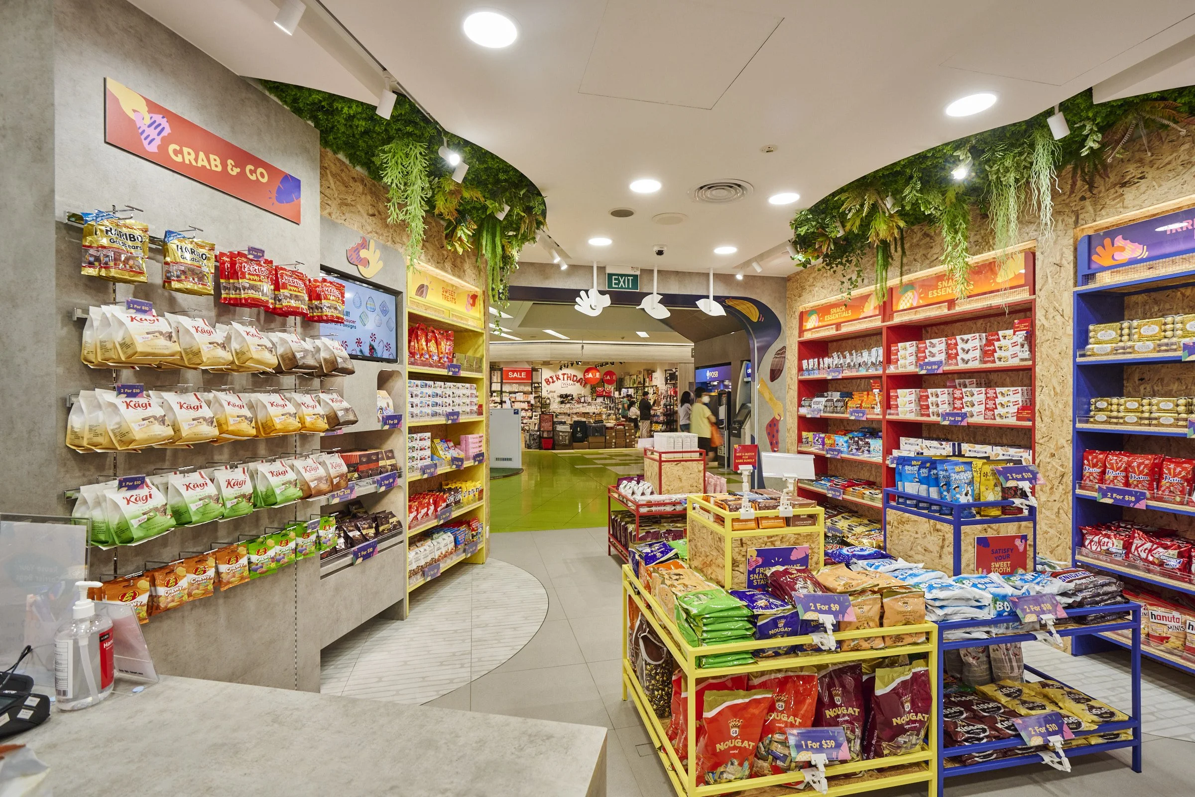 A retail store aisle with shelves stocked with snack foods, including chips, cookies, and candies, decorated with green plants along the ceiling and bright signage.