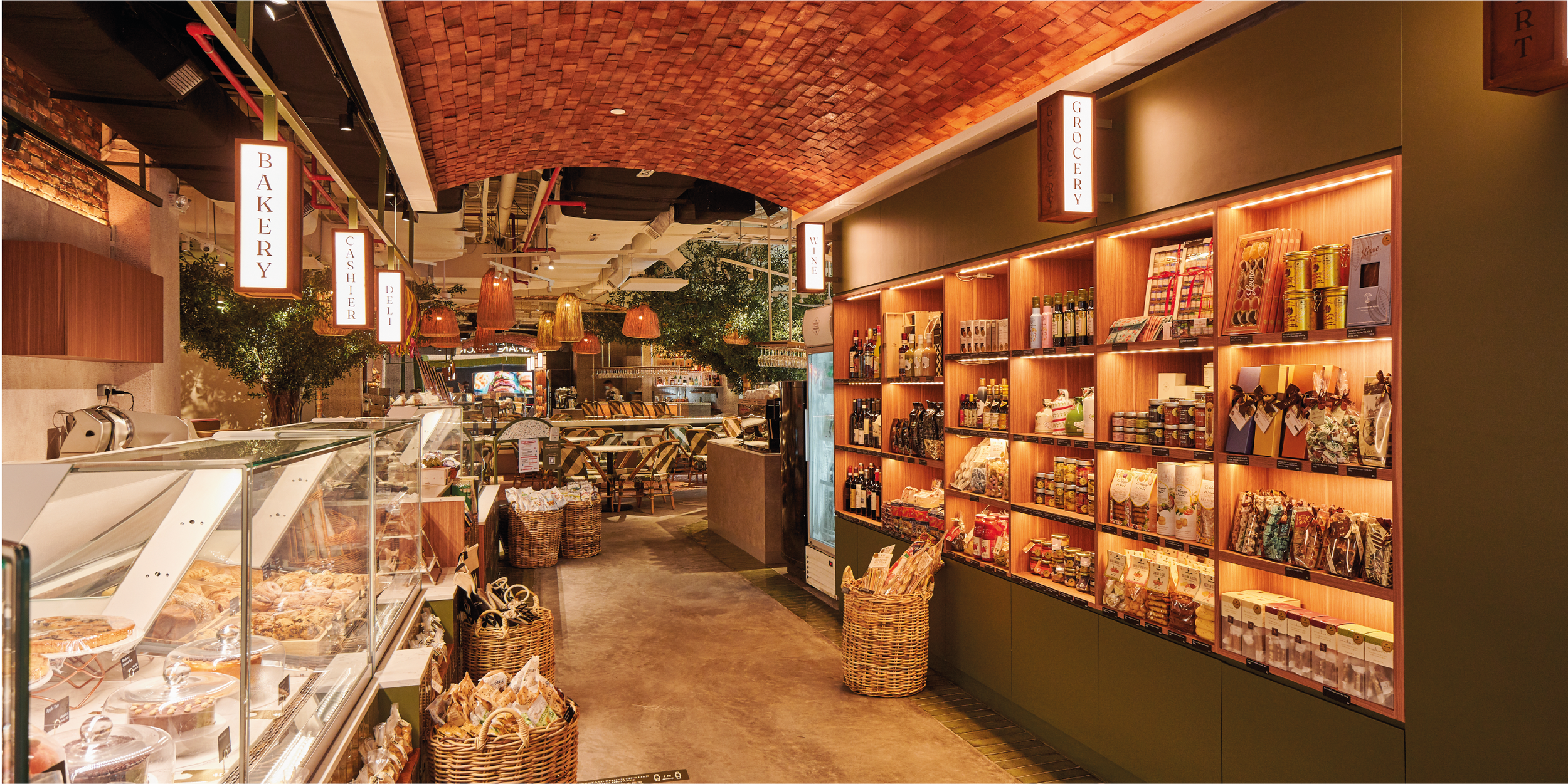 Interior of a grocery store with wooden shelving filled with food products, bakery section with baked goods in glass display cases, hanging pendant lights, and signs for bakery, cashier, deli, and grocery sections.