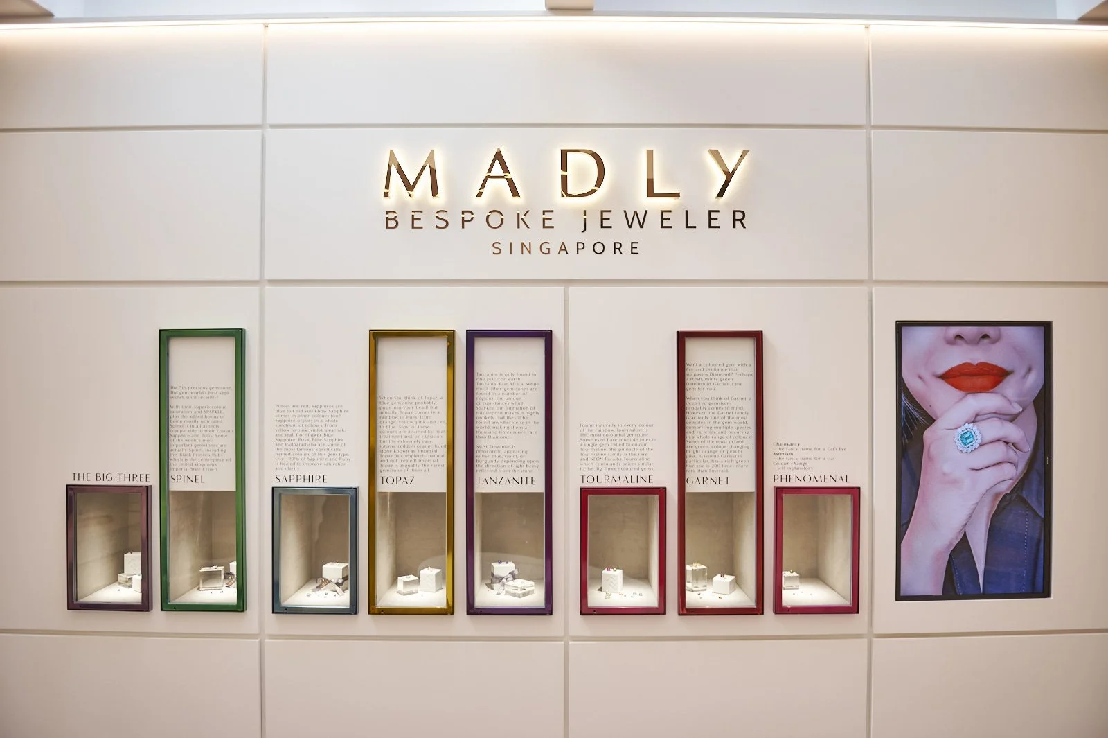 Display wall of MADLY Bespoke Jeweler in Singapore showcasing various gemstones including the big three, spinel, sapphire, topaz, tanzanite, tourmaline, garnet, and featuring a woman with her hand near her face wearing an elaborate ring.