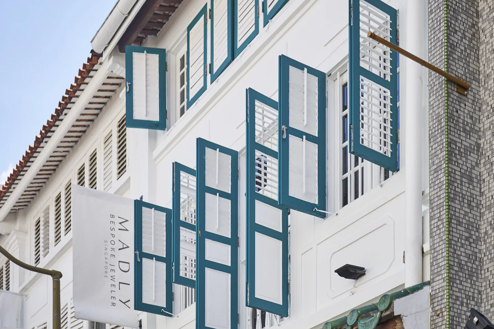 Exterior view of a white building with multiple blue shutters on the windows and a white flag that reads 'Madly' and 'Espoie Lewers Singafoe' hanging outside.