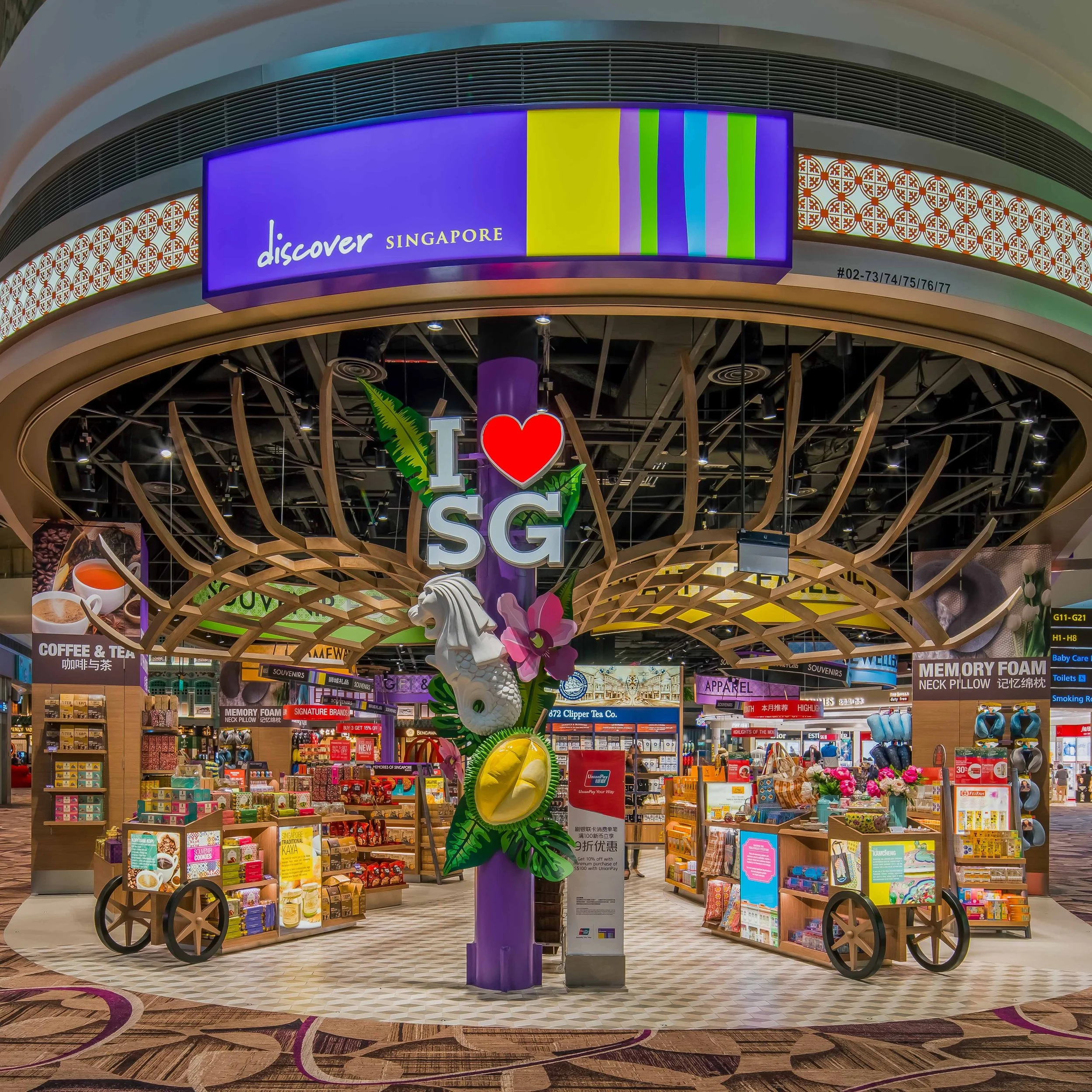 A retail store in Singapore with decorative signage and colorful displays of souvenirs, snacks, and travel items, including a prominent purple column with a sign that says 'I love SG' surrounded by tropical plants and flowers.