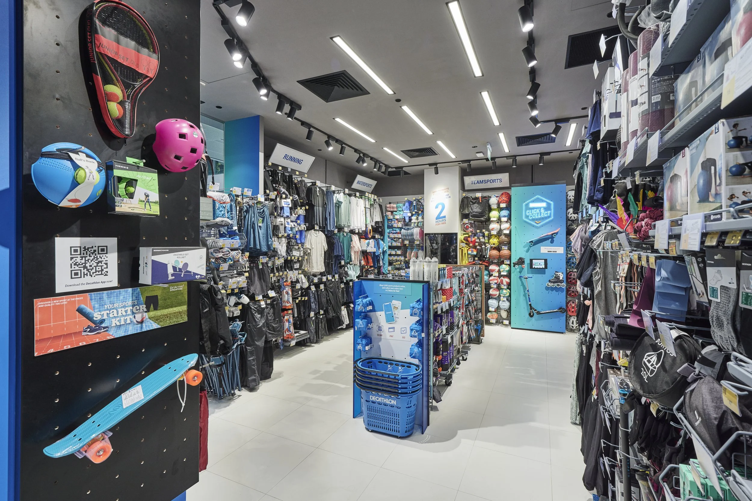 Interior of a sporting goods store with shelves stocked with athletic equipment, apparel, and accessories. Visible items include helmets, balls, skateboards, and clothing, with signs indicating sections like running, water sports, and team sports.