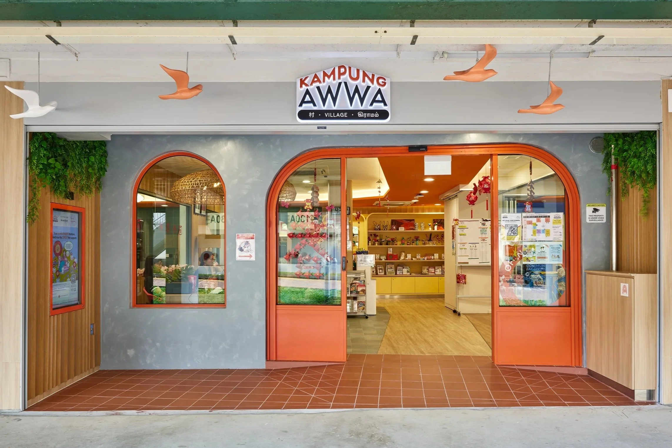 Entrance of a store called 'Kampung AWWA' with orange arched windows and door, greenery on sides, hanging bird decorations, and visible interior with yellow shelving and wall decorations.