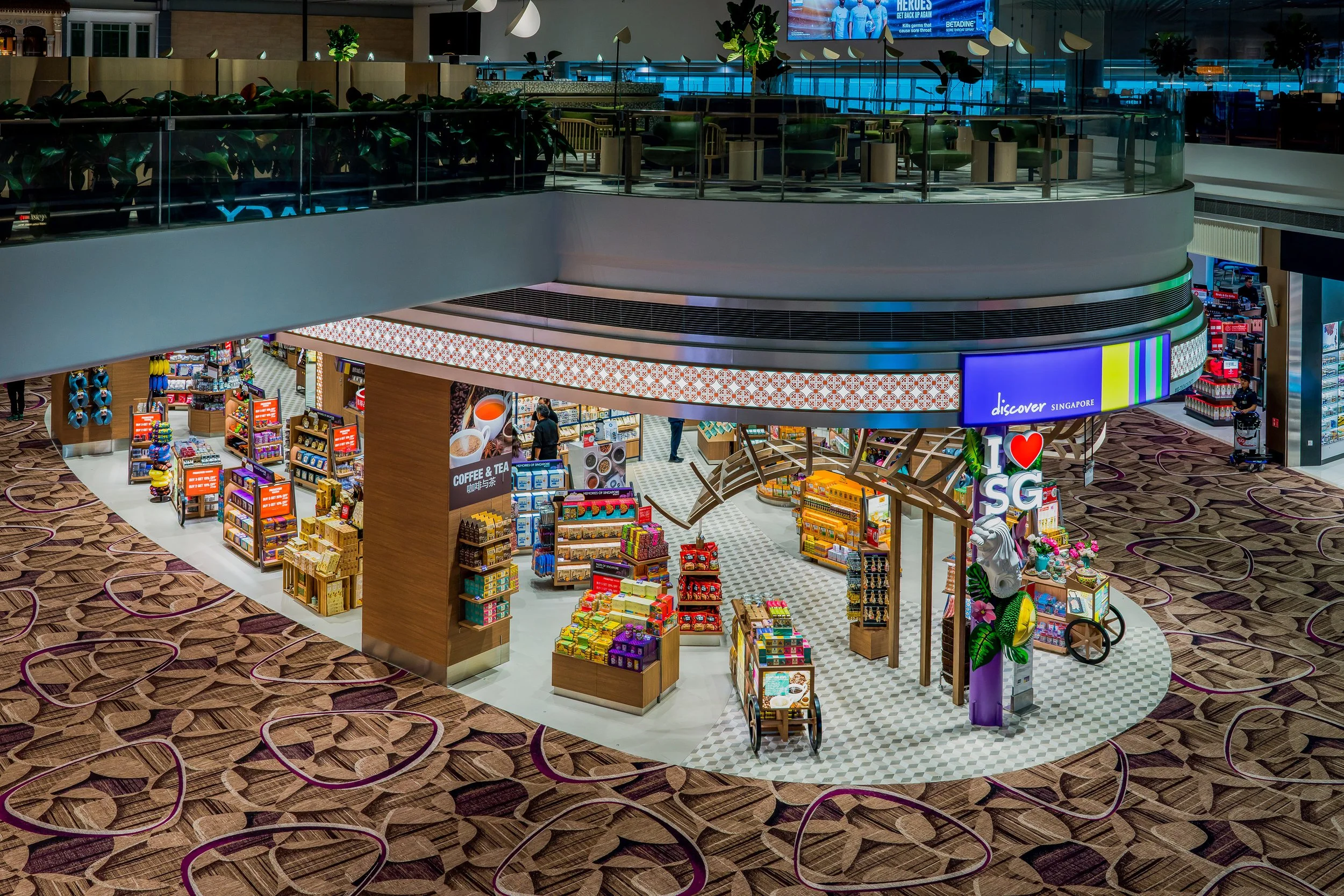 A multi-level shopping mall with a souvenir store on the lower level featuring colorful merchandise, a sign that says 'I love SG', and a patterned carpet with hearts.