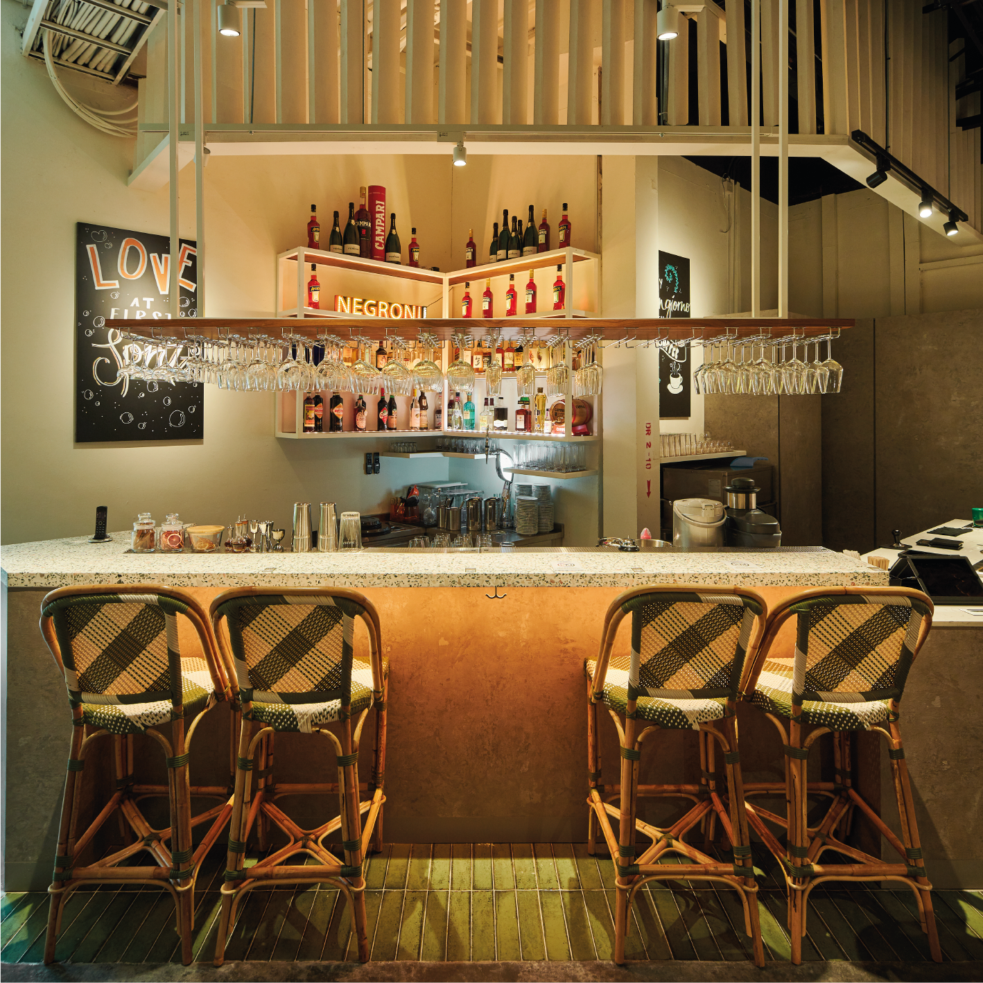 A stylish bar with a granite countertop, four woven rattan bar stools with green and beige cushions, hanging wine glasses, and shelves stocked with liquor bottles, with chalkboard signs and decorative lighting.