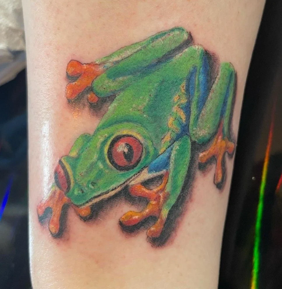 Colorful tattoo of a green tree frog with orange feet and red eyes on a person's arm.