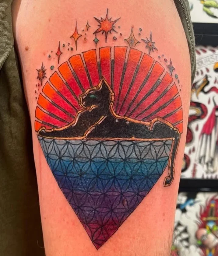 Colorful tattoo of a black cat sitting inside a diamond-shaped geometric pattern, with a vibrant red and orange sunburst behind it and star-like sparkles above.