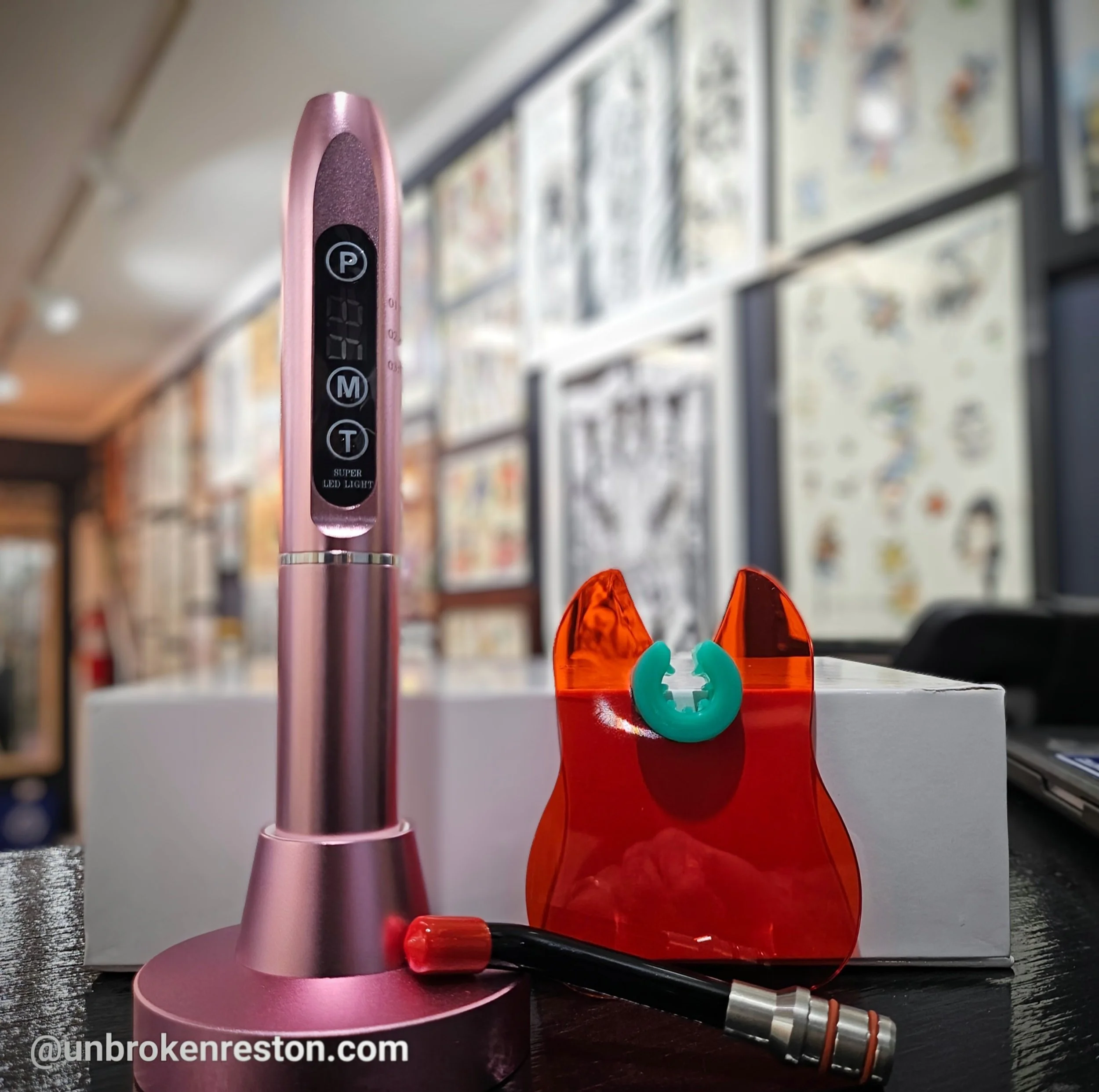 A pink LED facial treatment device, a red facial mask with a turquoise ring, and a black and red facial massage tool on a dark surface with framed artwork on the wall in the background.