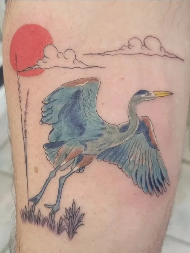 A tattoo of a heron standing in grass with a red sun and clouds in the background.