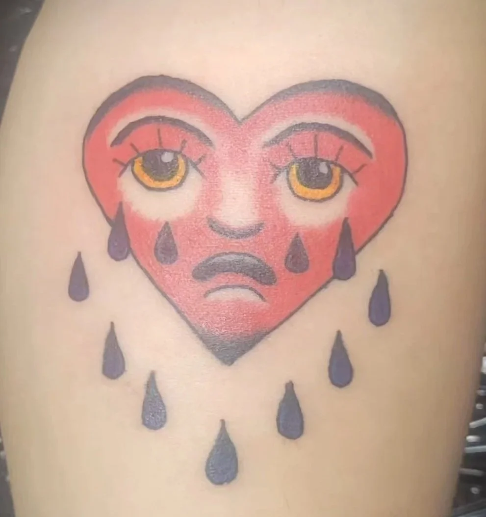 Tattoo of a red, sad heart with a human face, featuring large, expressive eyes and tears falling down.