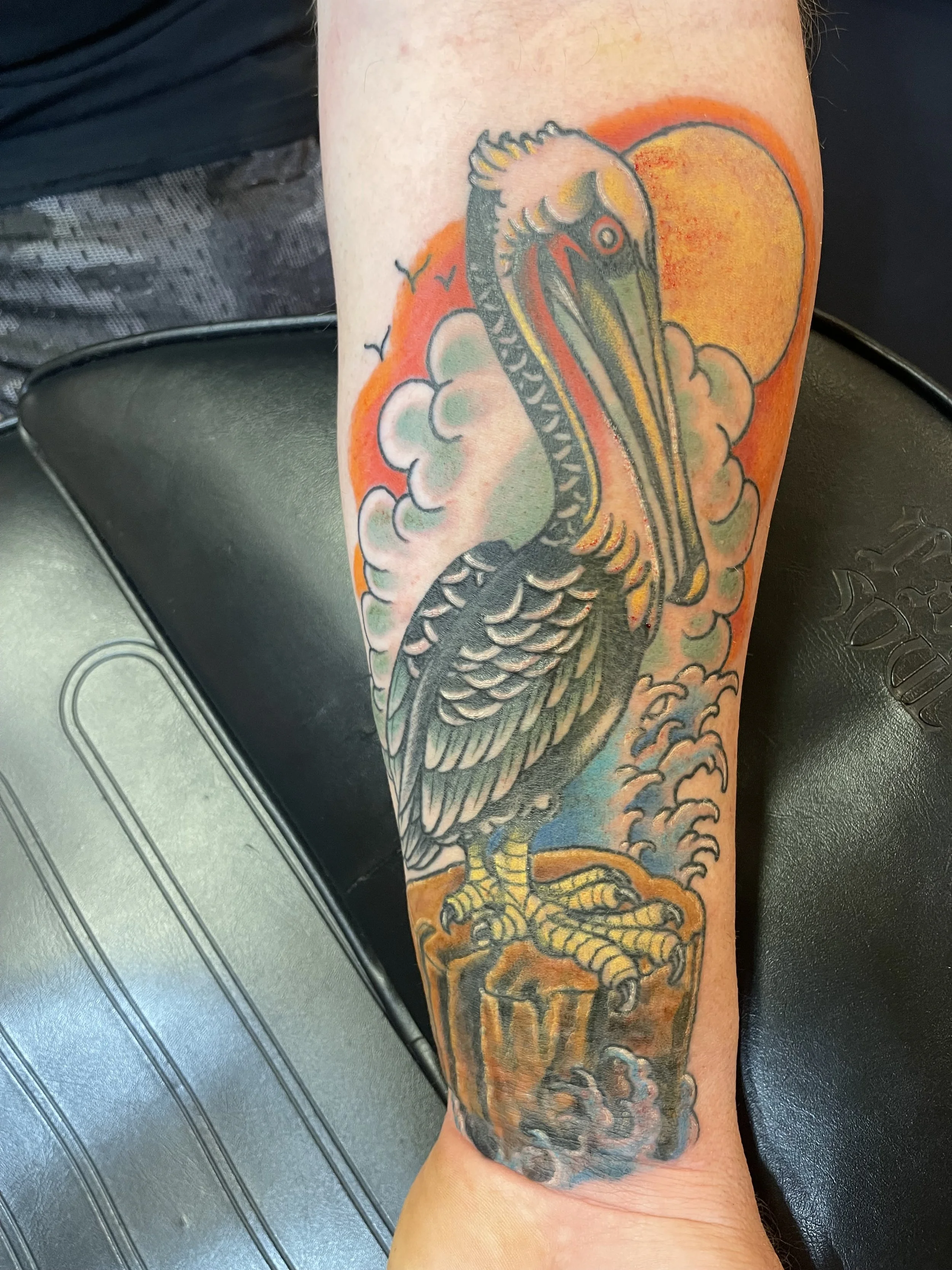Tattoo of a pelican perched on a post with clouds, a sun, and a blue sky background.