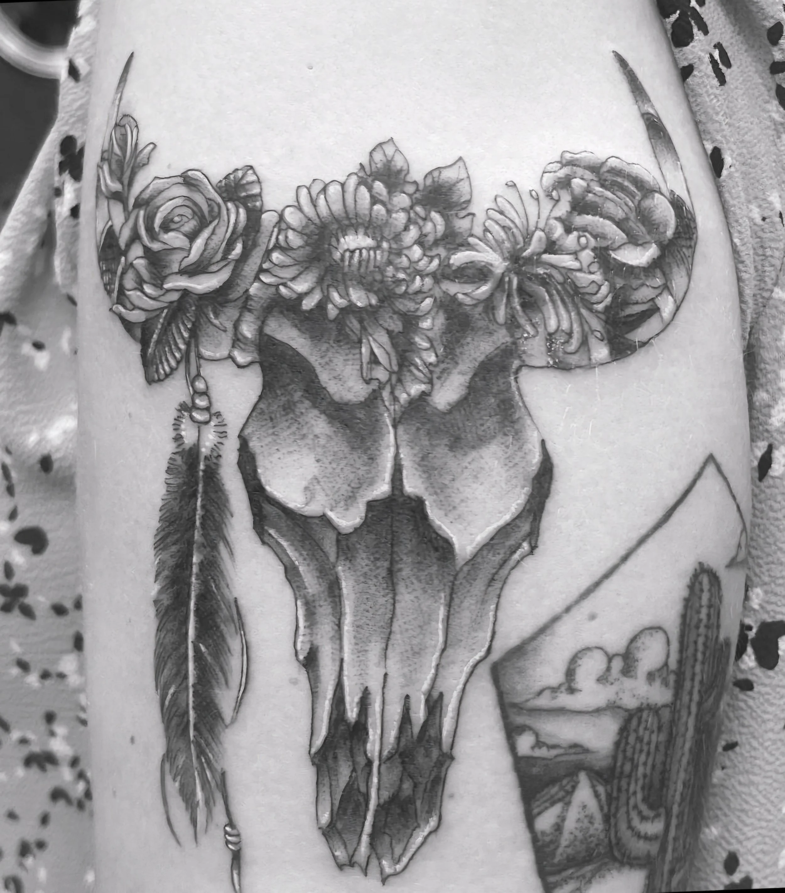 Tattoo of a cow skull with flowers on top, feathers on the sides, and desert landscape with cacti in the background.