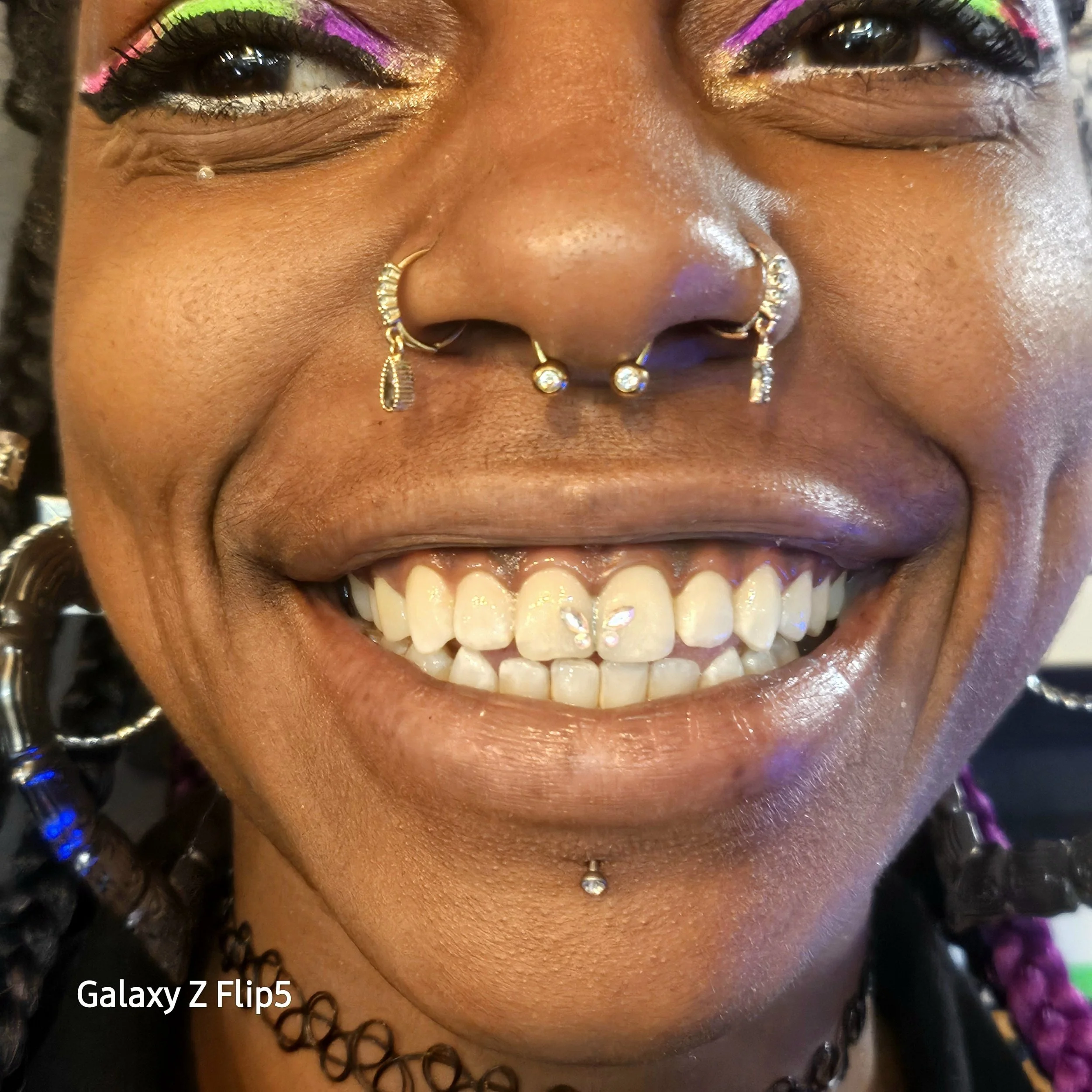 Close-up of a person smiling with colorful eye makeup, multiple facial piercings, and a choker necklace.