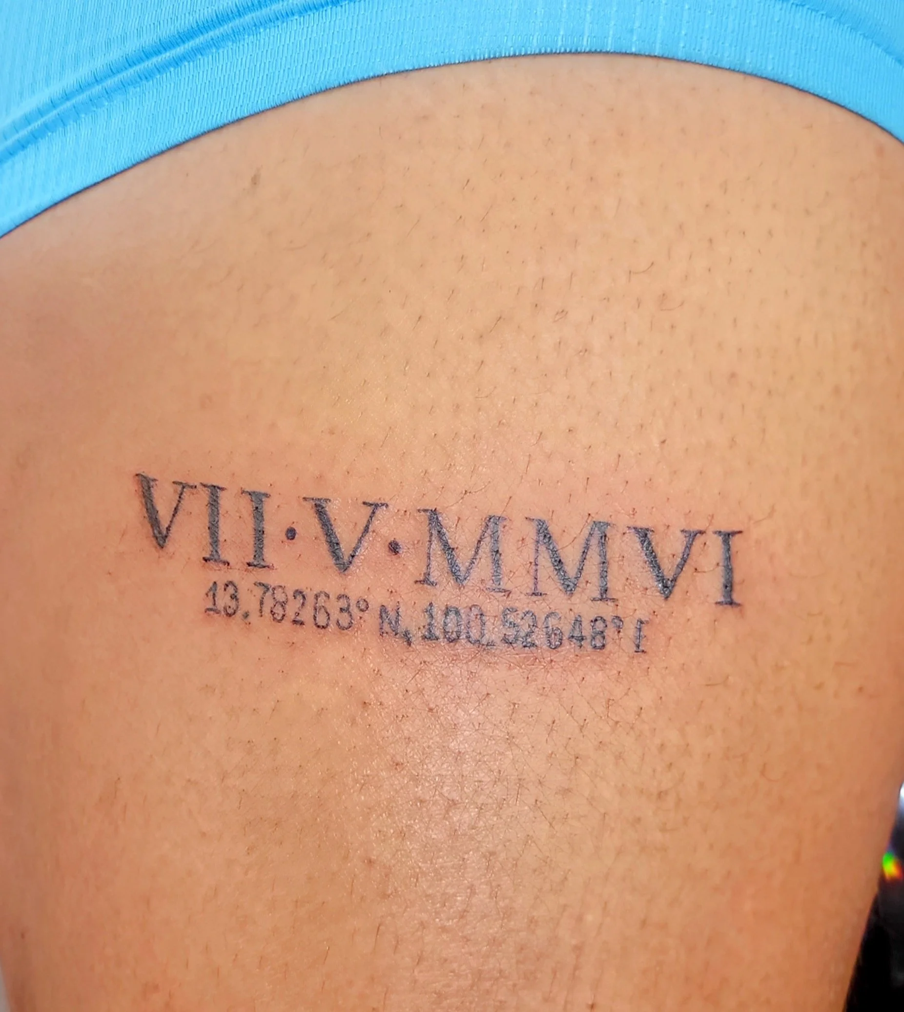 Tattoo on person's skin with Roman numerals, coordinates, and a degree symbol, partially covered by a blue fabric.
