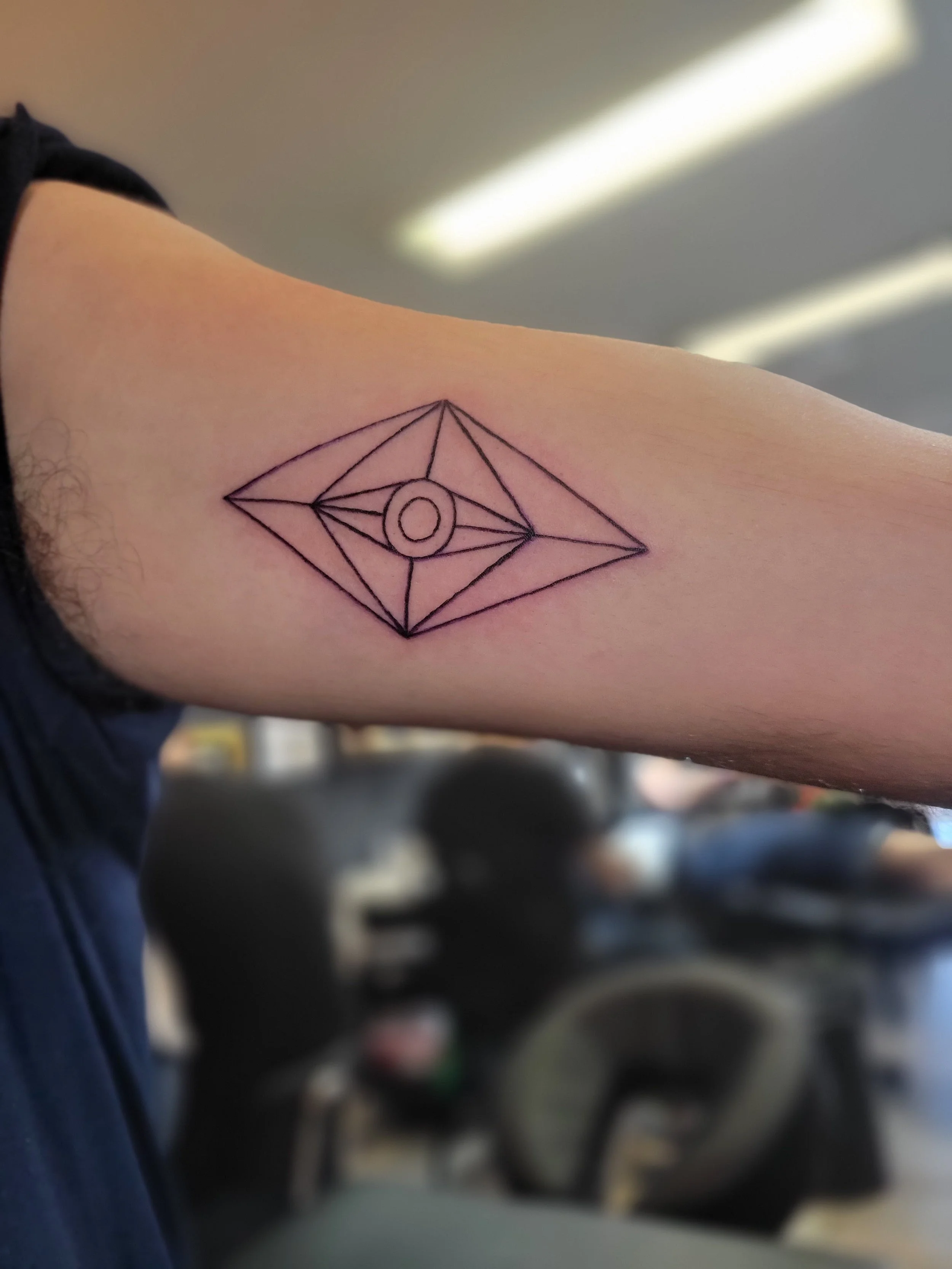 A geometric tattoo on a person's upper arm featuring a diamond shape with multiple intersecting lines and a circle in the center.