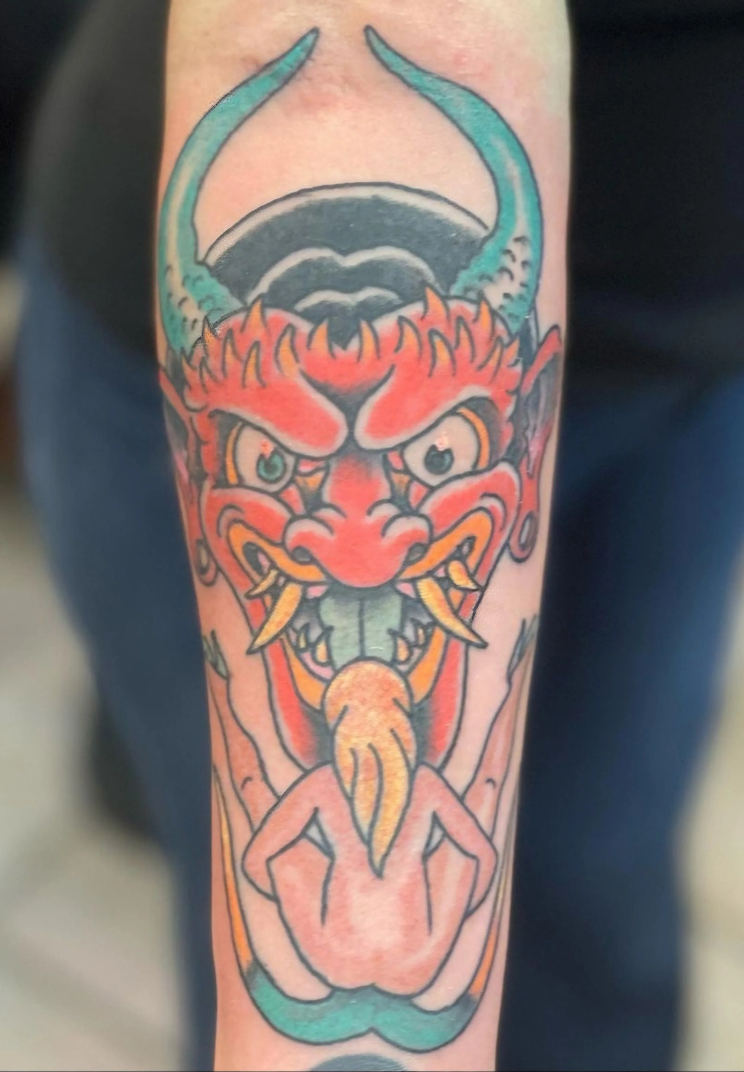 Colorful Japanese-style tattoo of a fierce red dragon with blue horns, sharp teeth, and a long green tongue, shown on a person's arm.