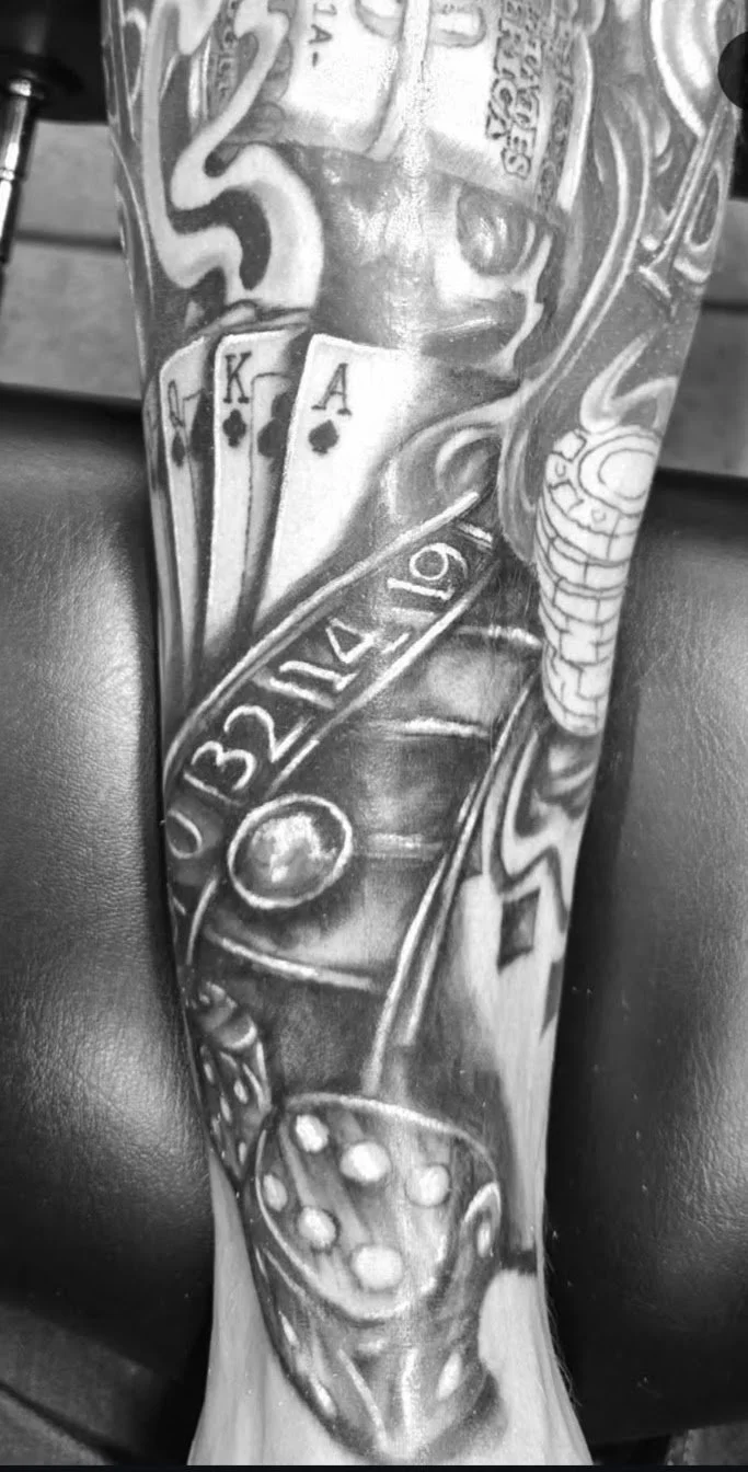 Close-up of a tattooed arm featuring playing cards, poker chips, dice, and dollar bills in black and white.