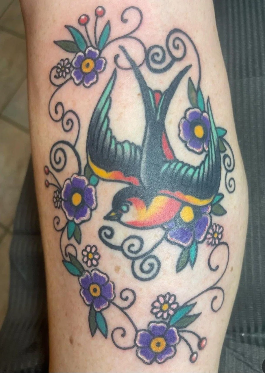 Tattoo of a colorful bird surrounded by purple flowers and floral swirls.
