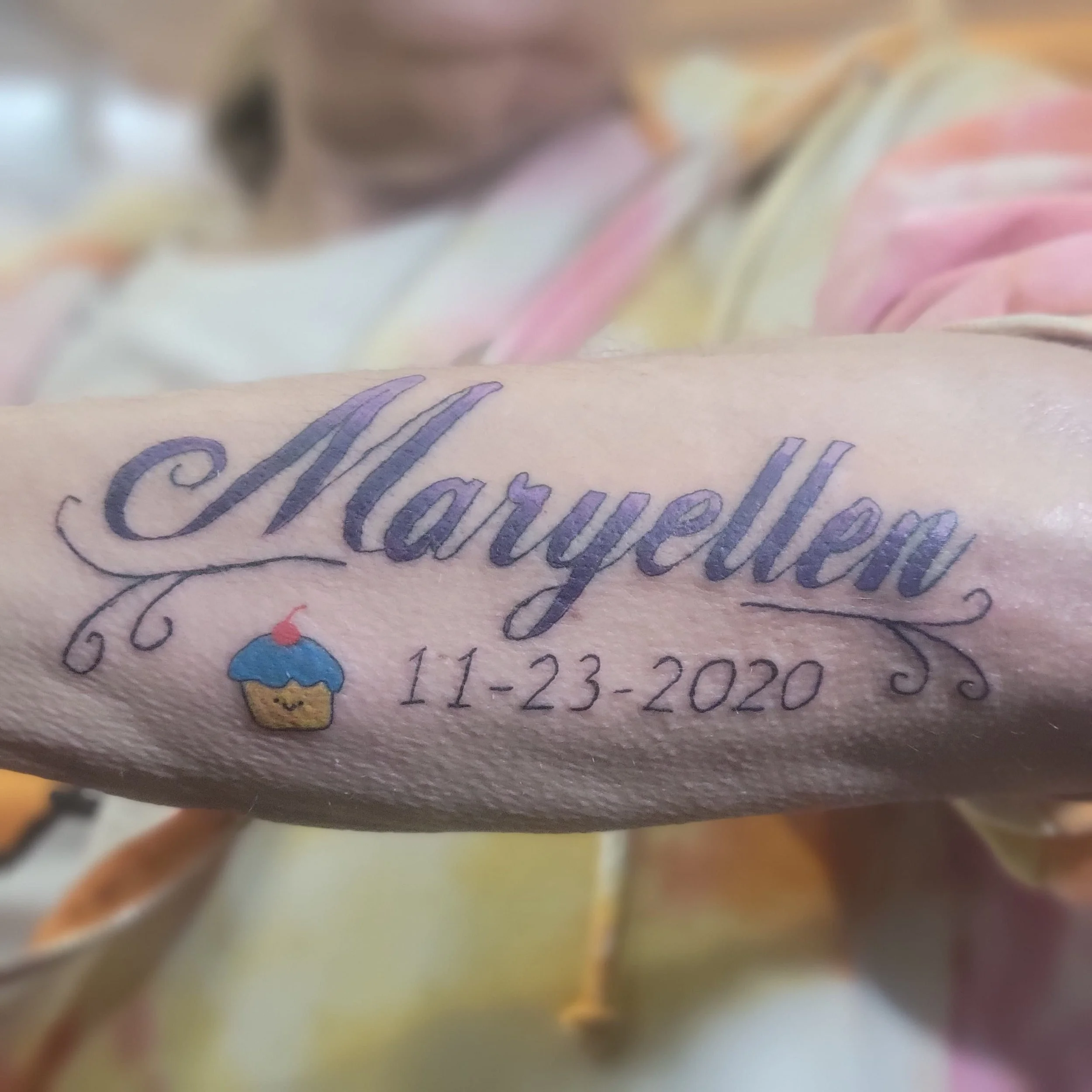 Tattoo of the name "Maryellen" in cursive with the date "11-23-2020" and a small cupcake with blue frosting and a cherry on top.