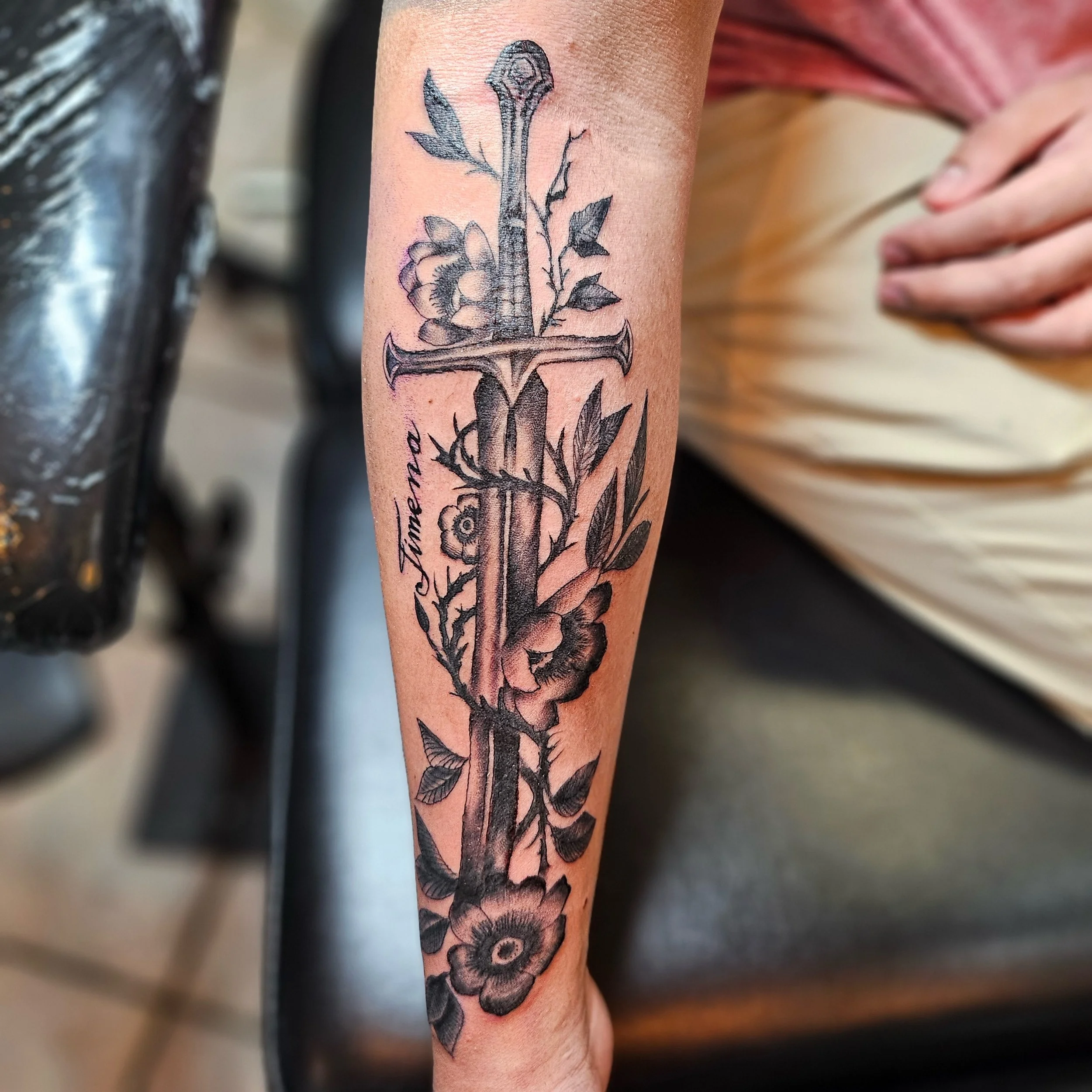 Black and gray tattoo of a sword entwined with flowers and leaves on a person's forearm.