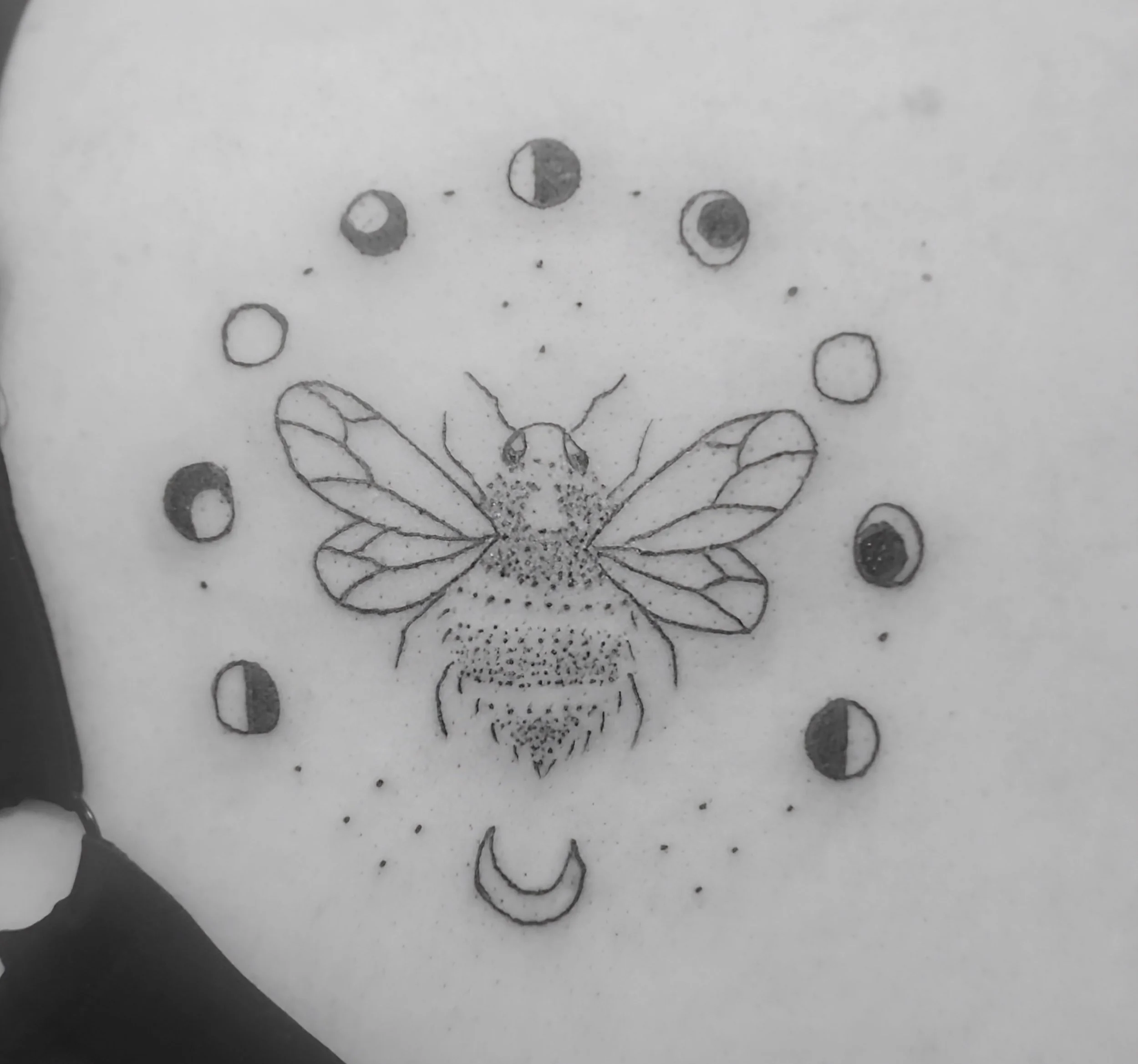 Tattoo of a bee with phases of the moon and a crescent moon below it, surrounded by small dots.
