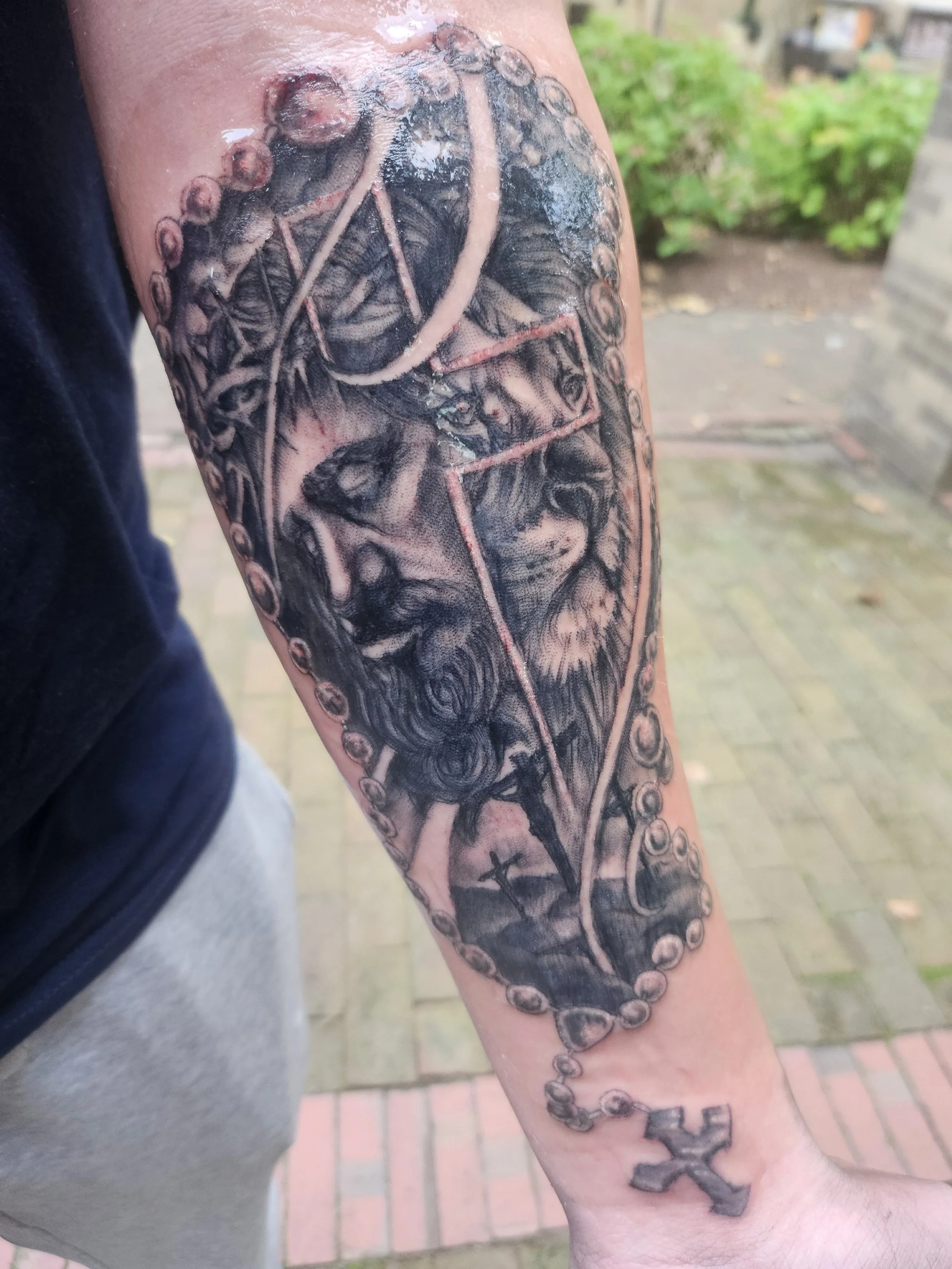 Black and gray tattoo depicting a lion, a portrait of a woman, and a rosary with a cross on the person's arm.