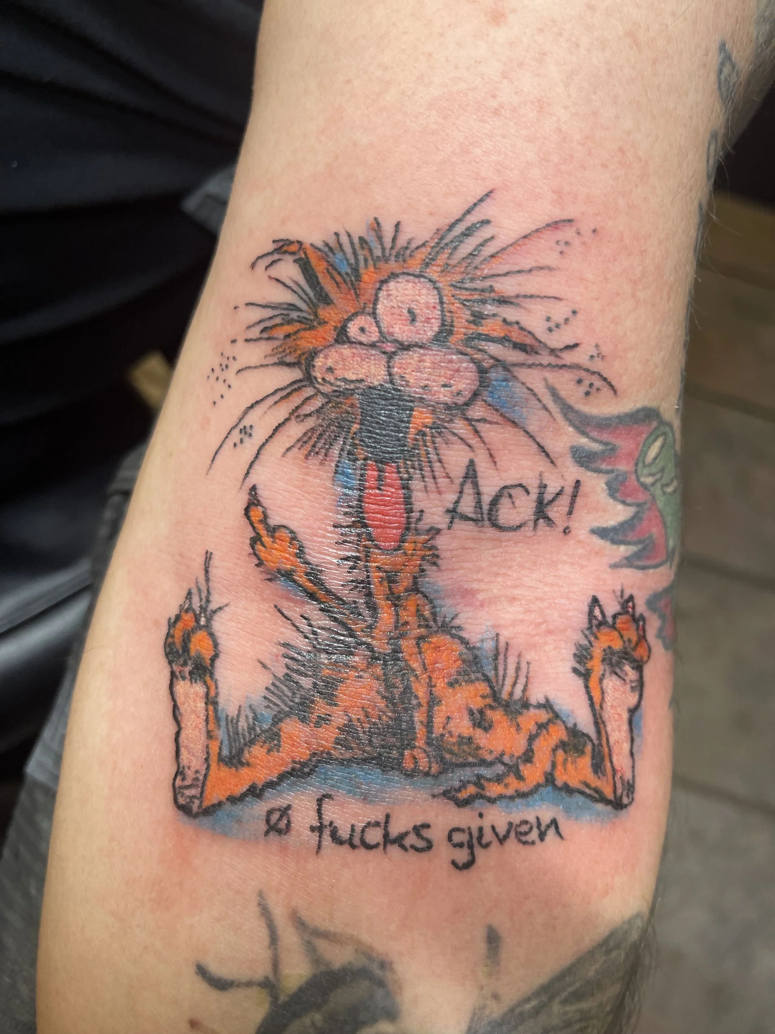 A colorful tattoo of a cartoon cat with wild hair, sticking out its tongue, with the word 'BACK!' next to it. The cat is lying on its back with its paws upwards, and the phrase 'a fucks given' is written below.
