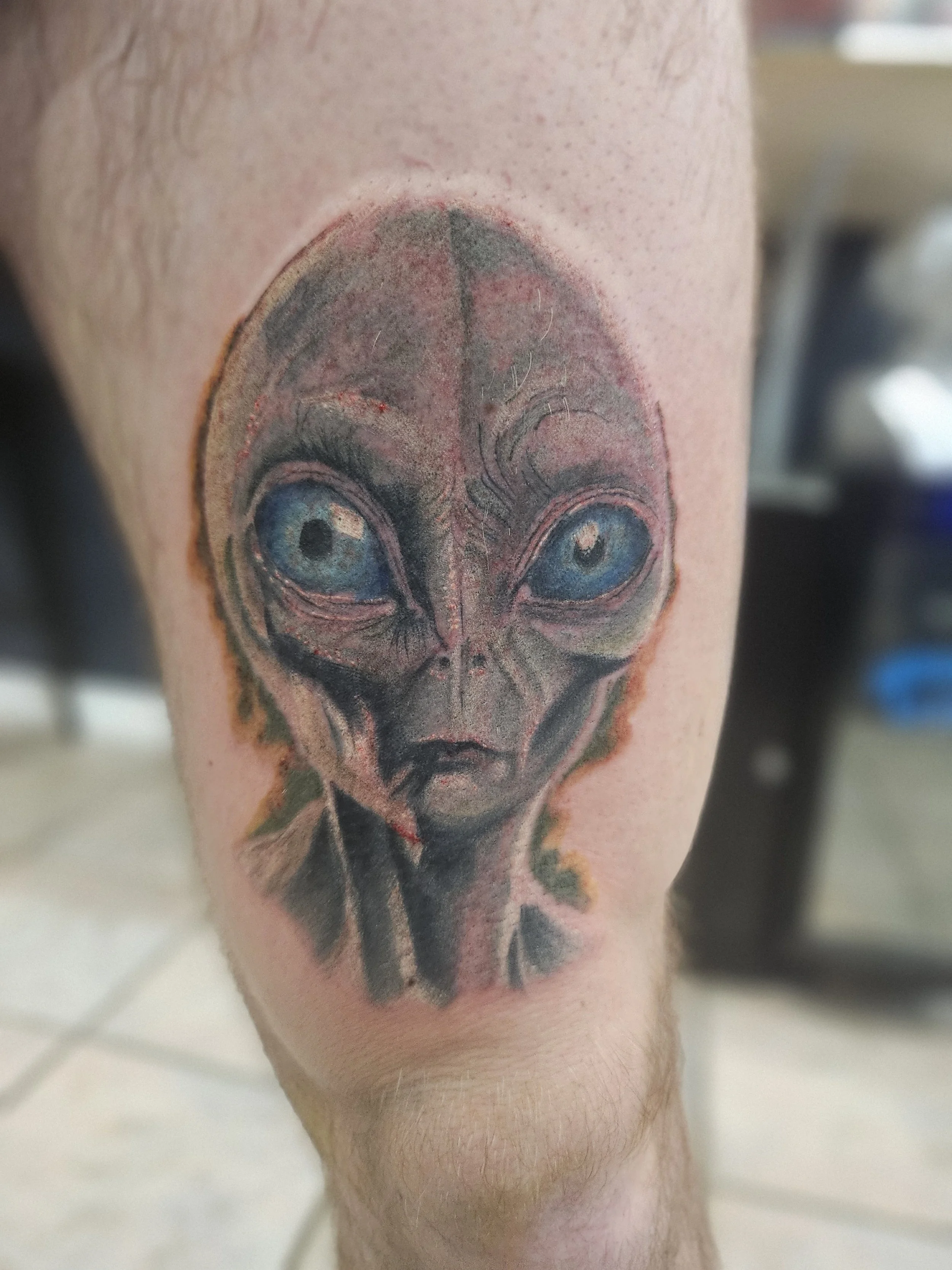 Tattoo of an alien's face with large eyes and elongated head on a person's upper arm.