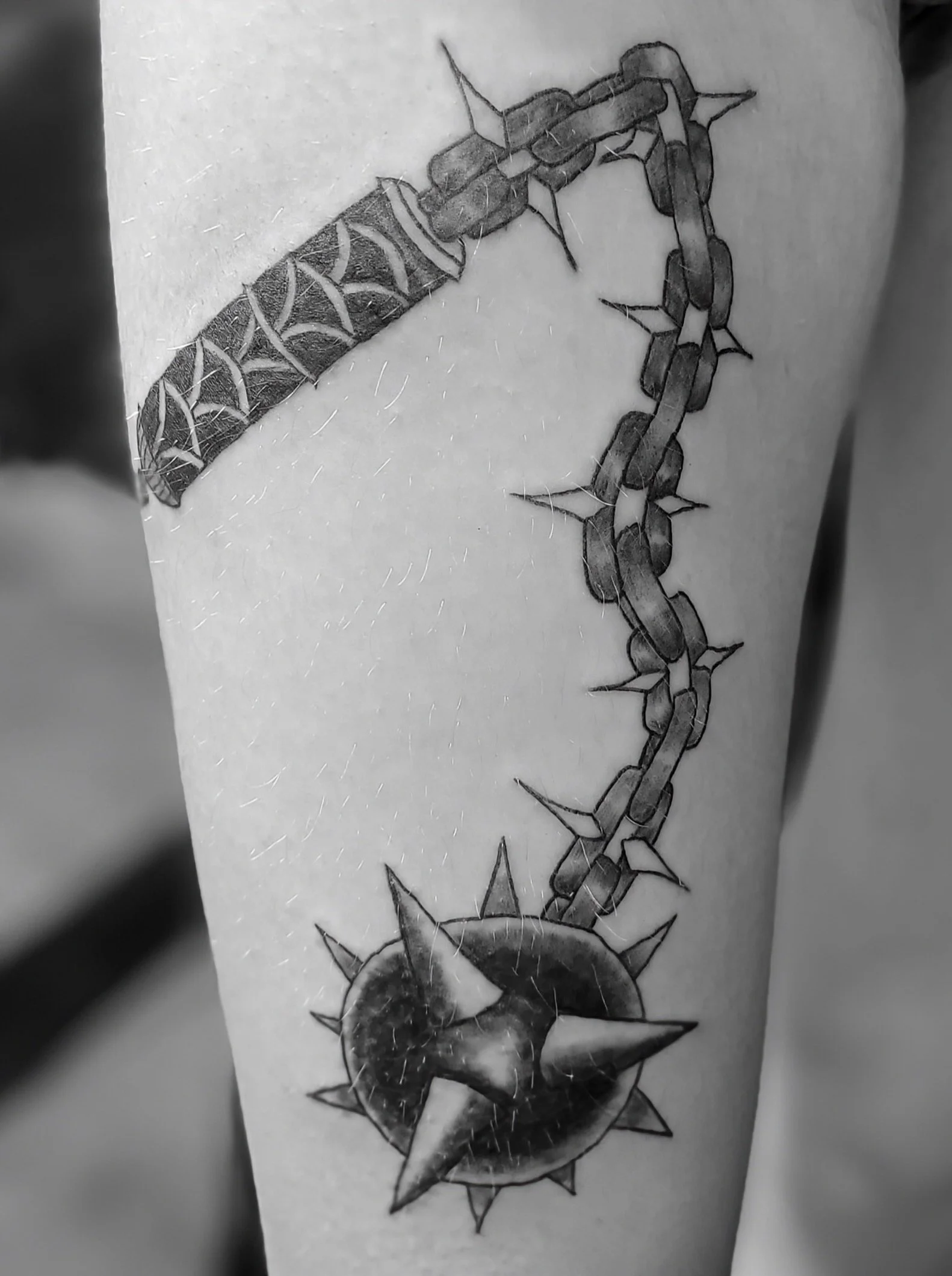 Black and white tattoo of a shuriken with spikes and a chain attached, on a person's arm.