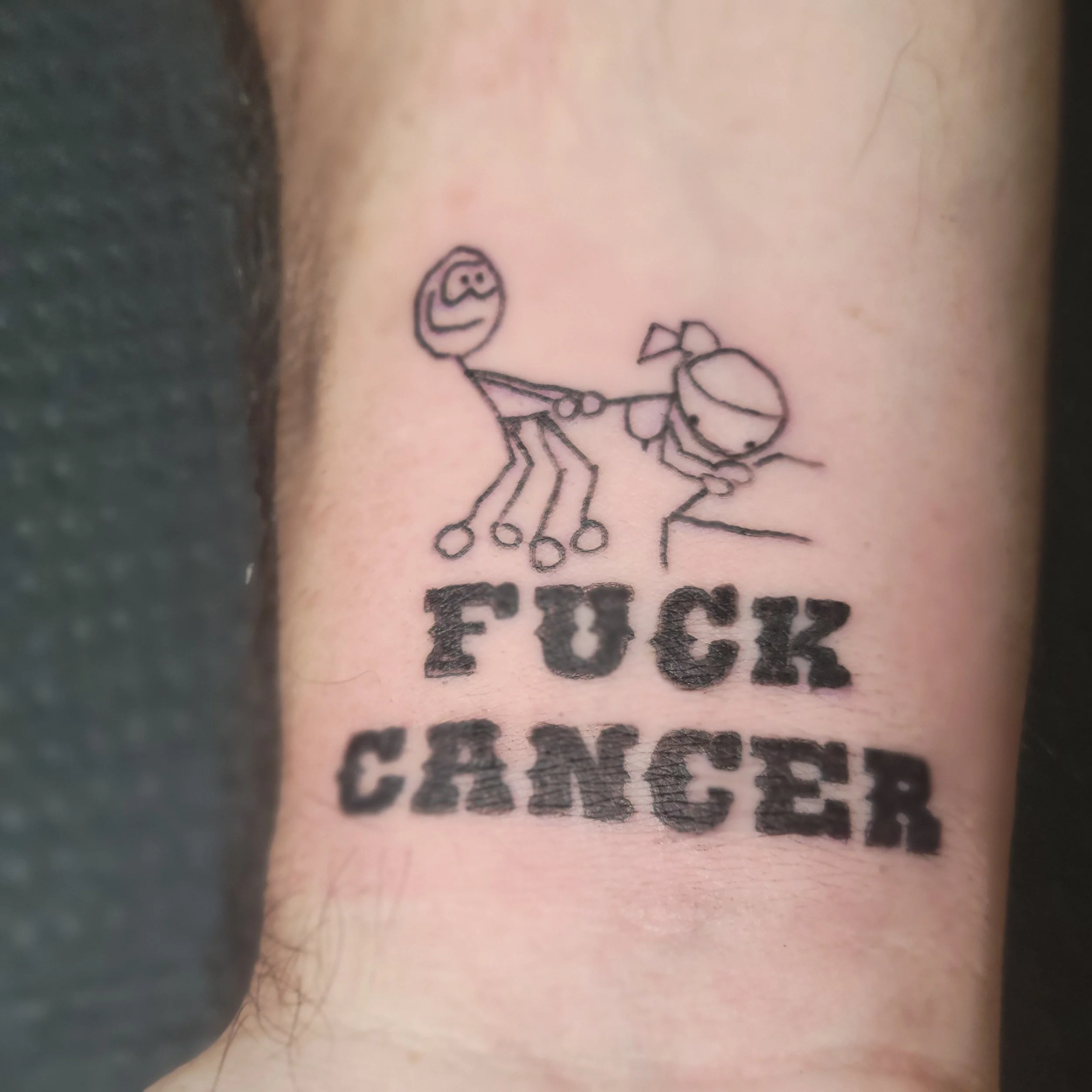 Tattoo of a stick figure girl with a ponytail holding a balloon that has a sad face, and the words 'FUCK CANCER' in bold black letters.