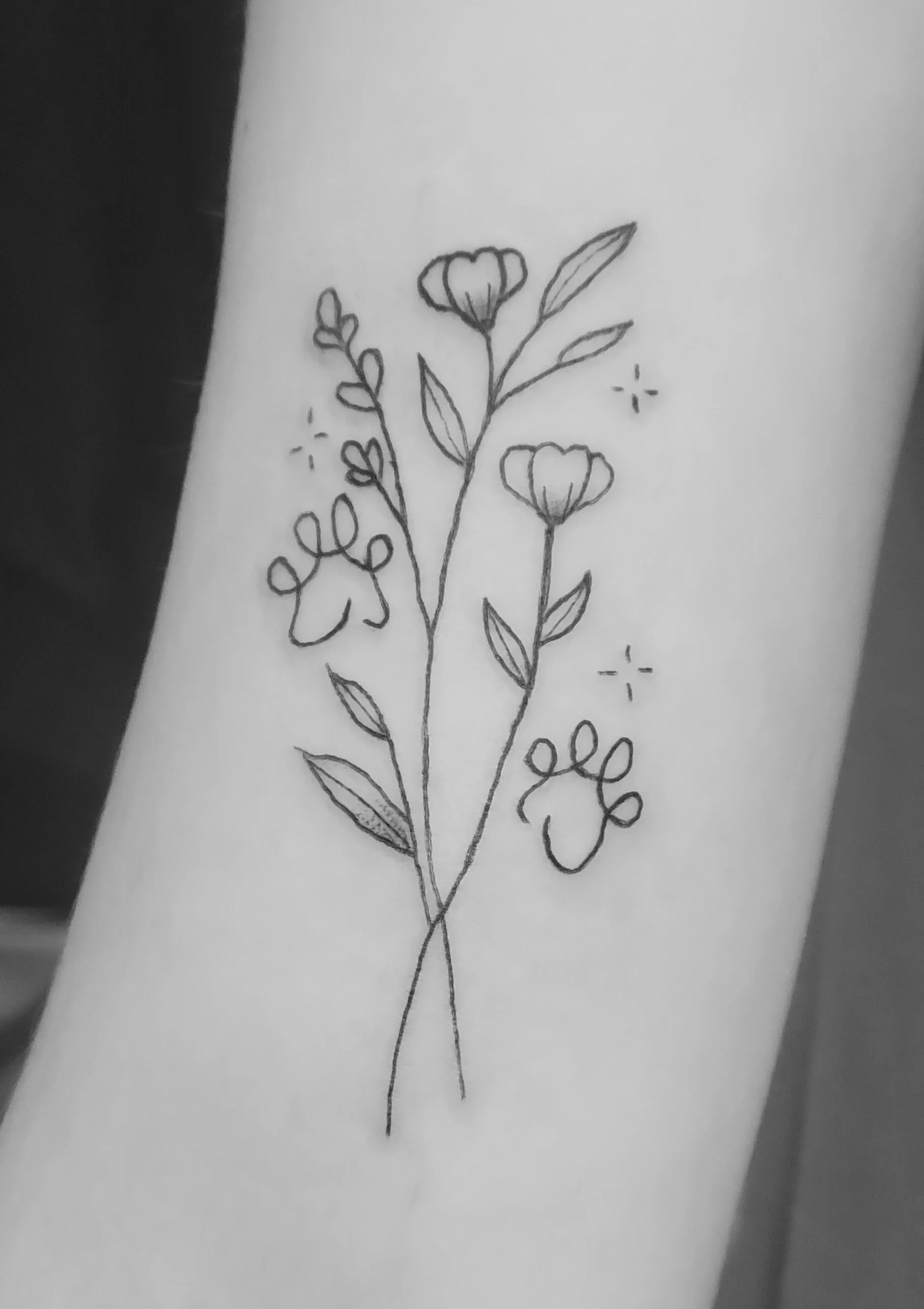 Black and white photo of a line drawing of a flower bouquet tattoo on someone’s skin, featuring two large flowers, leaves, and small decorative elements.