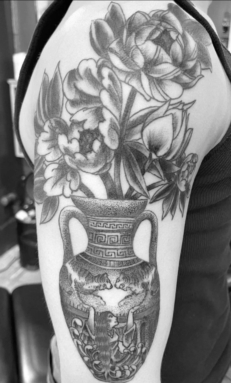 Black and gray tattoo of a vase with flowers and a woman in a decorative environment on a person's upper arm.