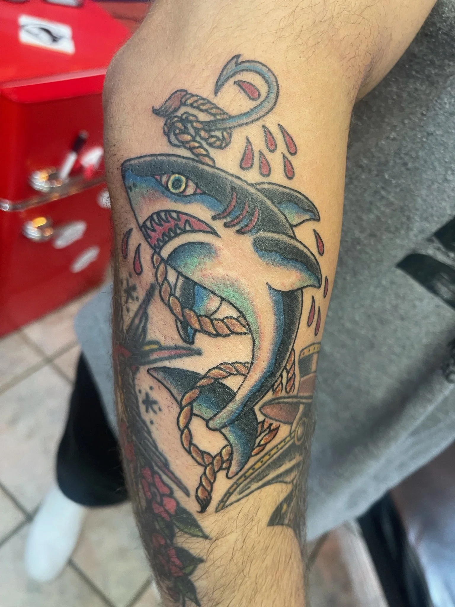 Tattoo of a shark with a rope around its body, tears, and a fishing hook in its mouth on a person's arm.