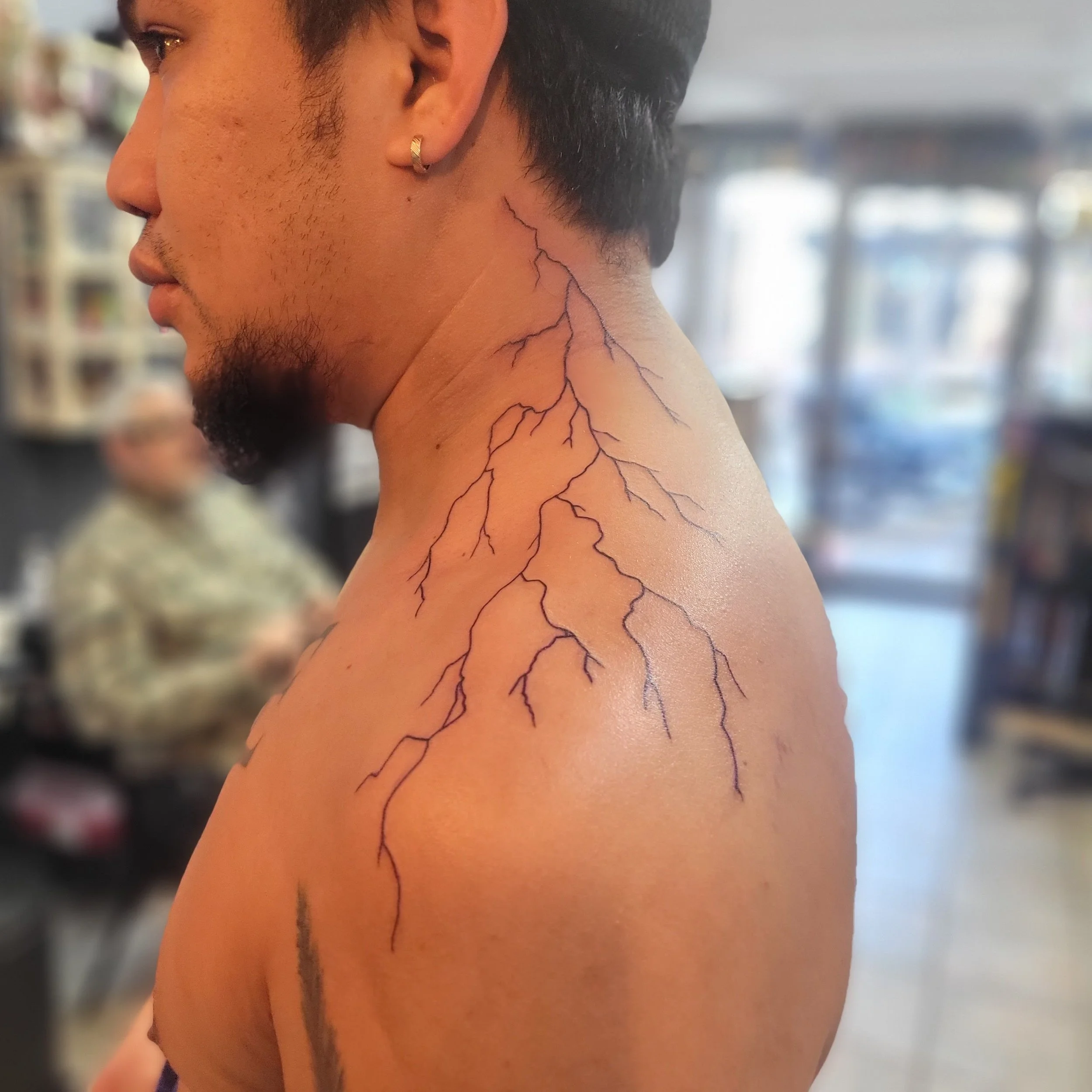A man with short dark hair and a beard, wearing a small hoop earring, has a tattoo of lightning bolts on his neck and shoulder.