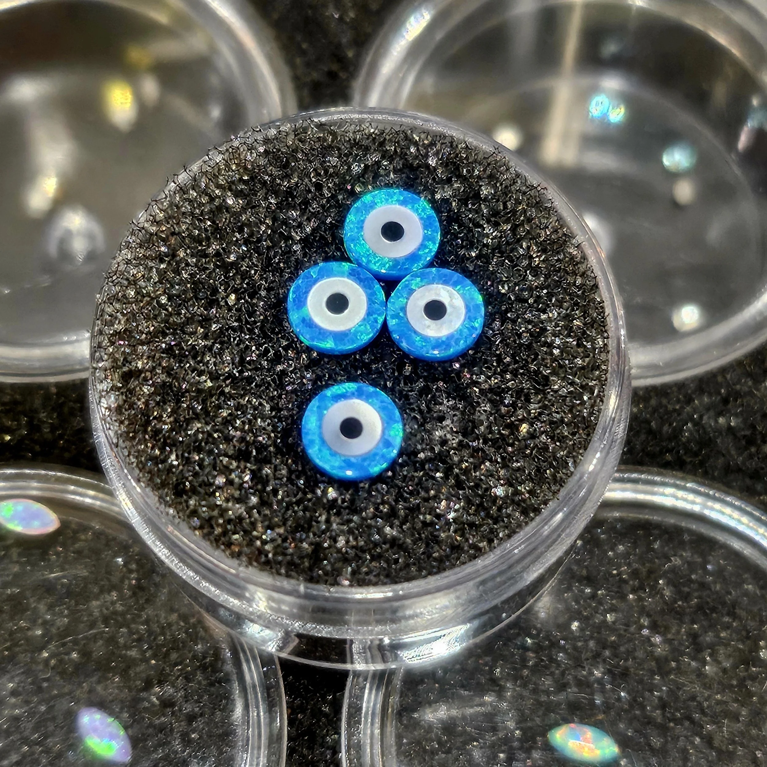 Close-up of four blue and white circle beads with black centers inside a clear plastic container, surrounded by other similar containers.