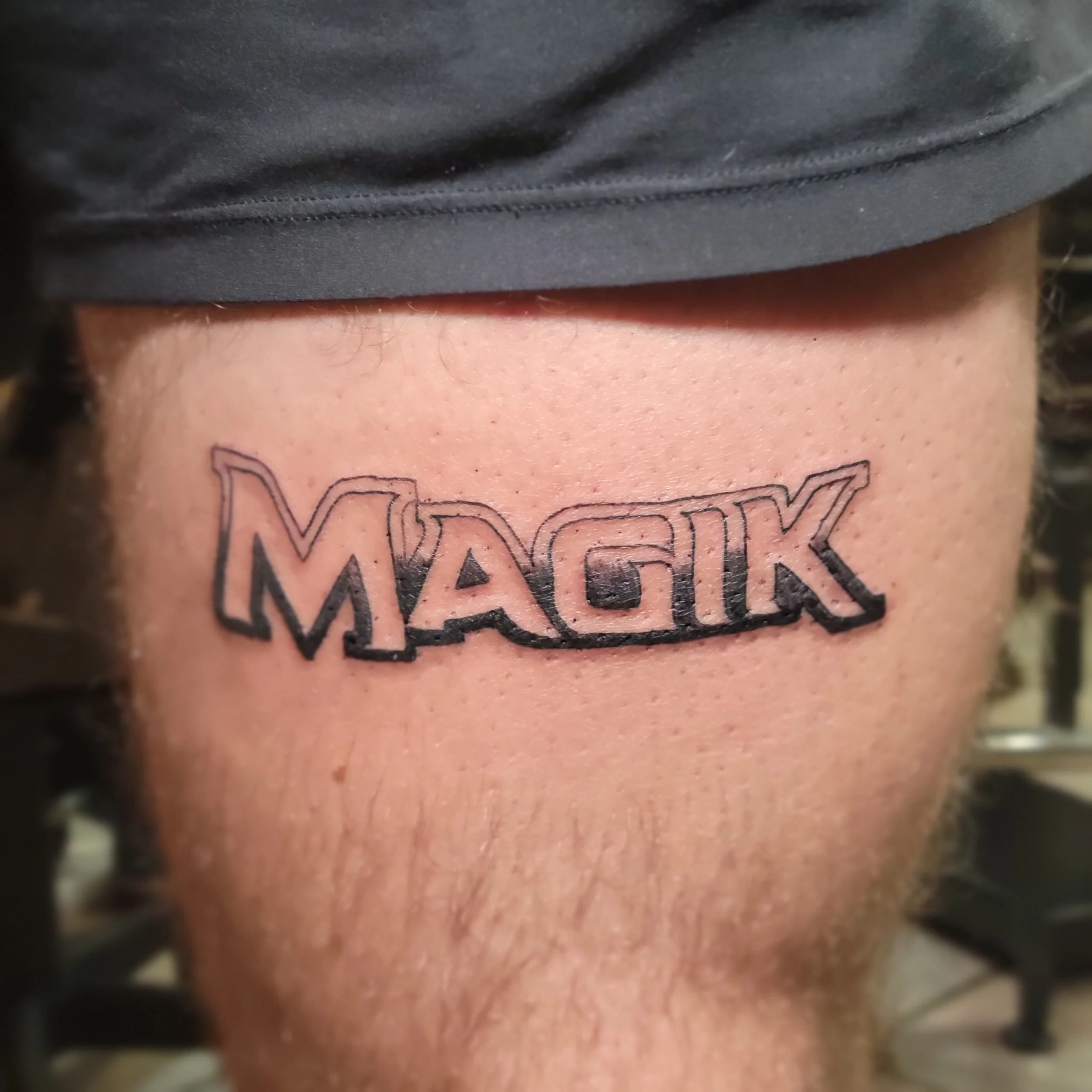 Close-up of a freshly done tattoo reading 'MAGIK' in bold, stylized black ink on a person's thigh.