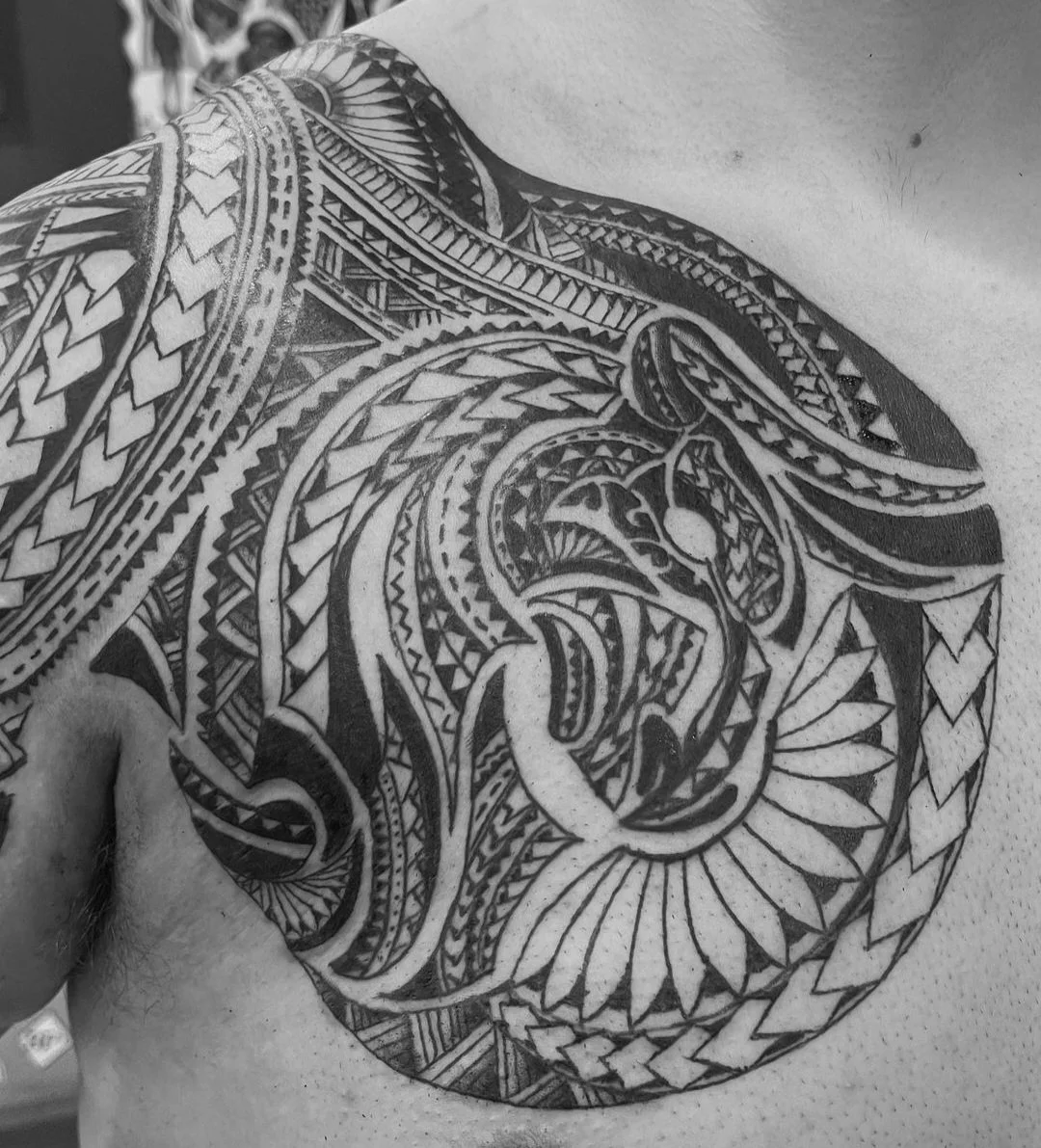 Black and white tribal-style tattoo on a person's shoulder and chest area, featuring intricate patterns and detailed line work.