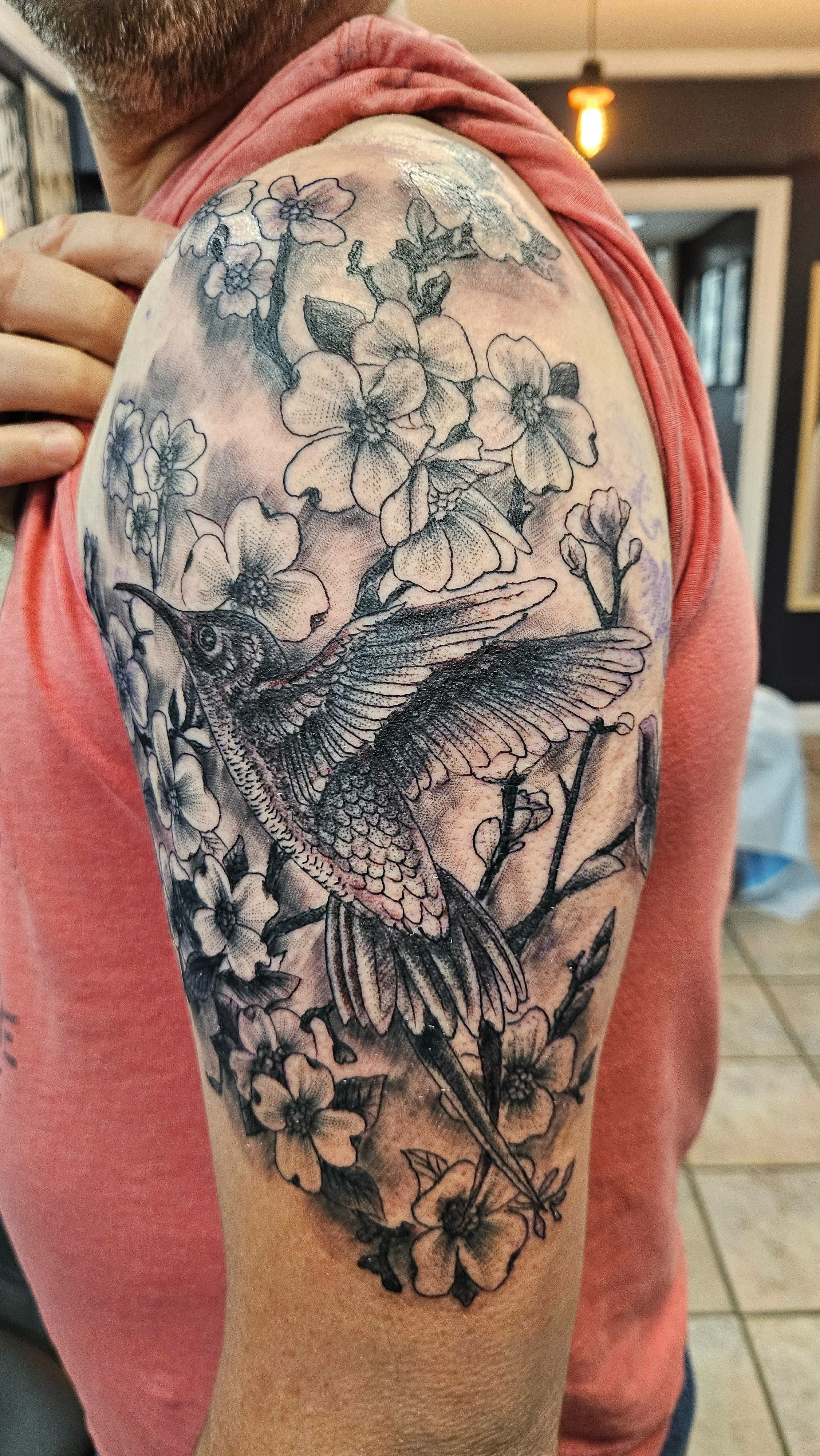 Tattoo of a hummingbird with outstretched wings surrounded by cherry blossoms on person's upper arm.