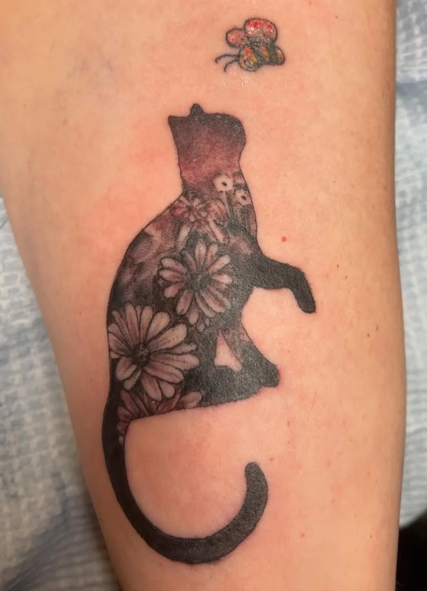 Tattoo of a cat silhouette filled with floral patterns, with a bee hovering above it.